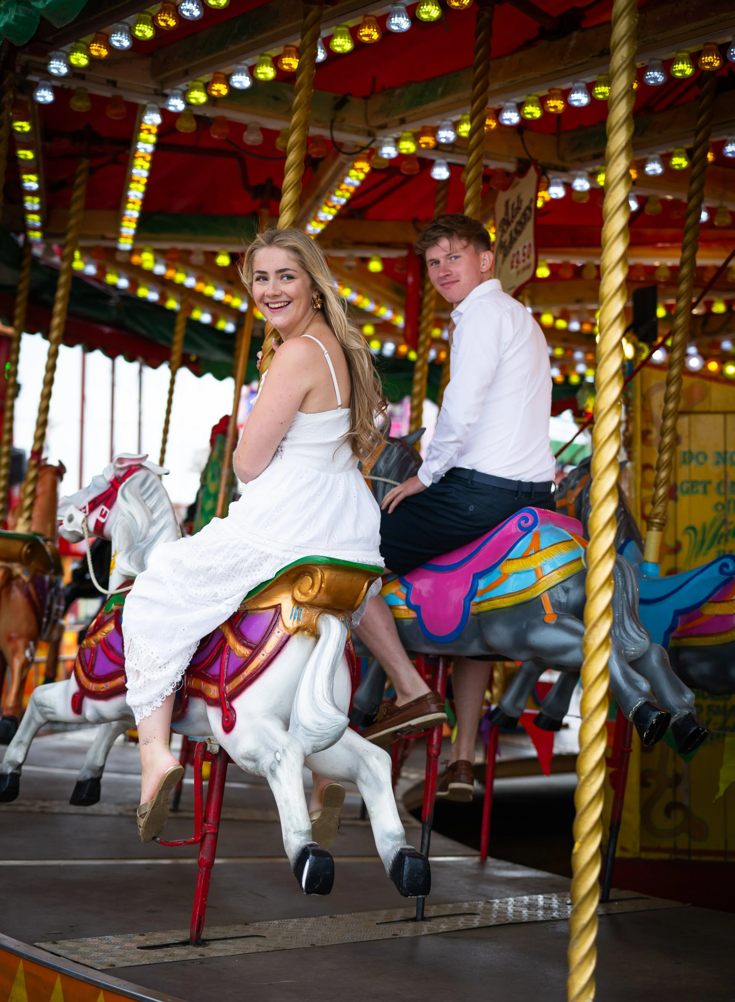 Young woman in white dress and young man in white shirt riding a carousel horse at an amusement park, with colorful lights in the background.