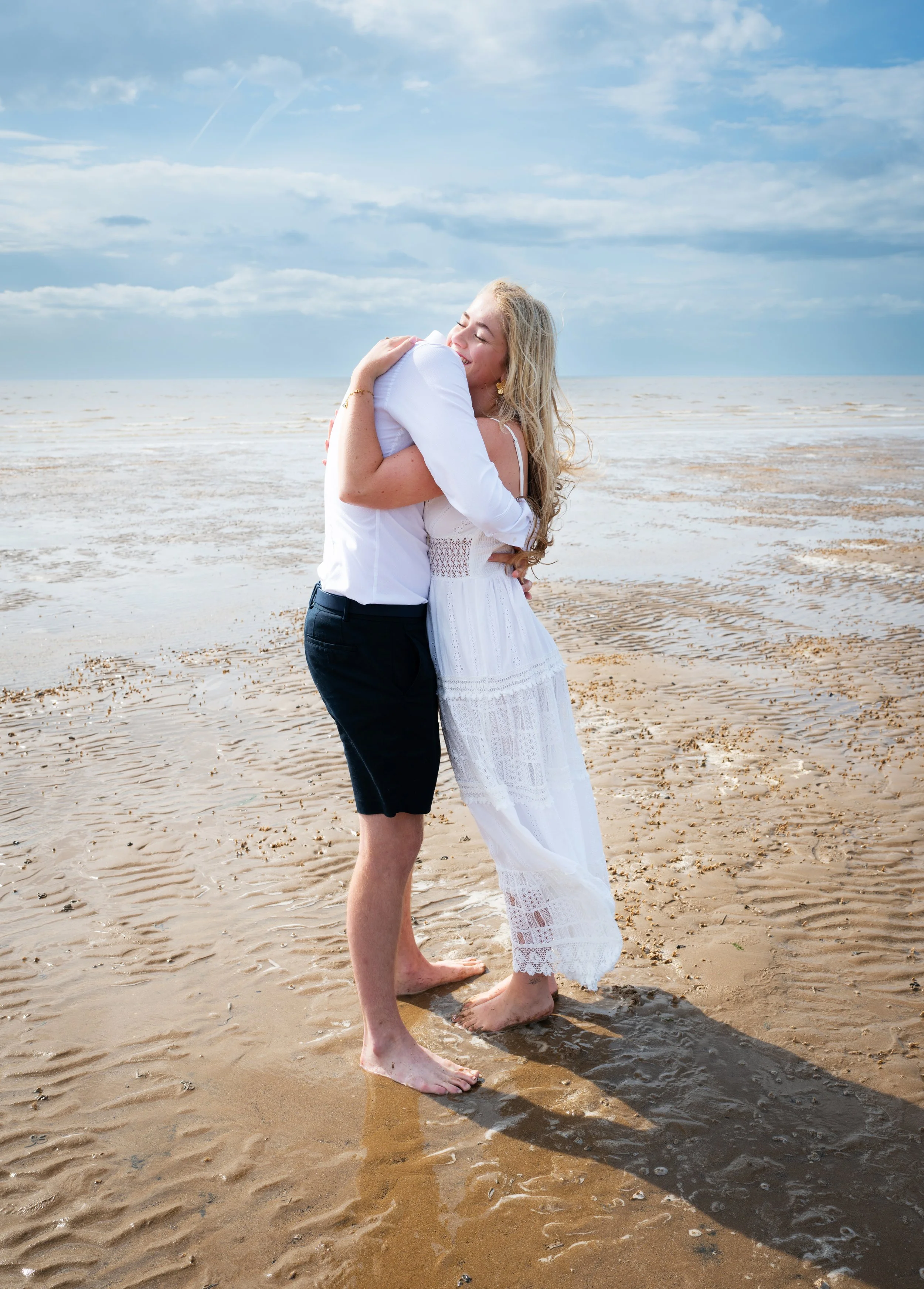 A couple hugging on a sandy beach, standing barefoot with the ocean and partly cloudy sky in the background.