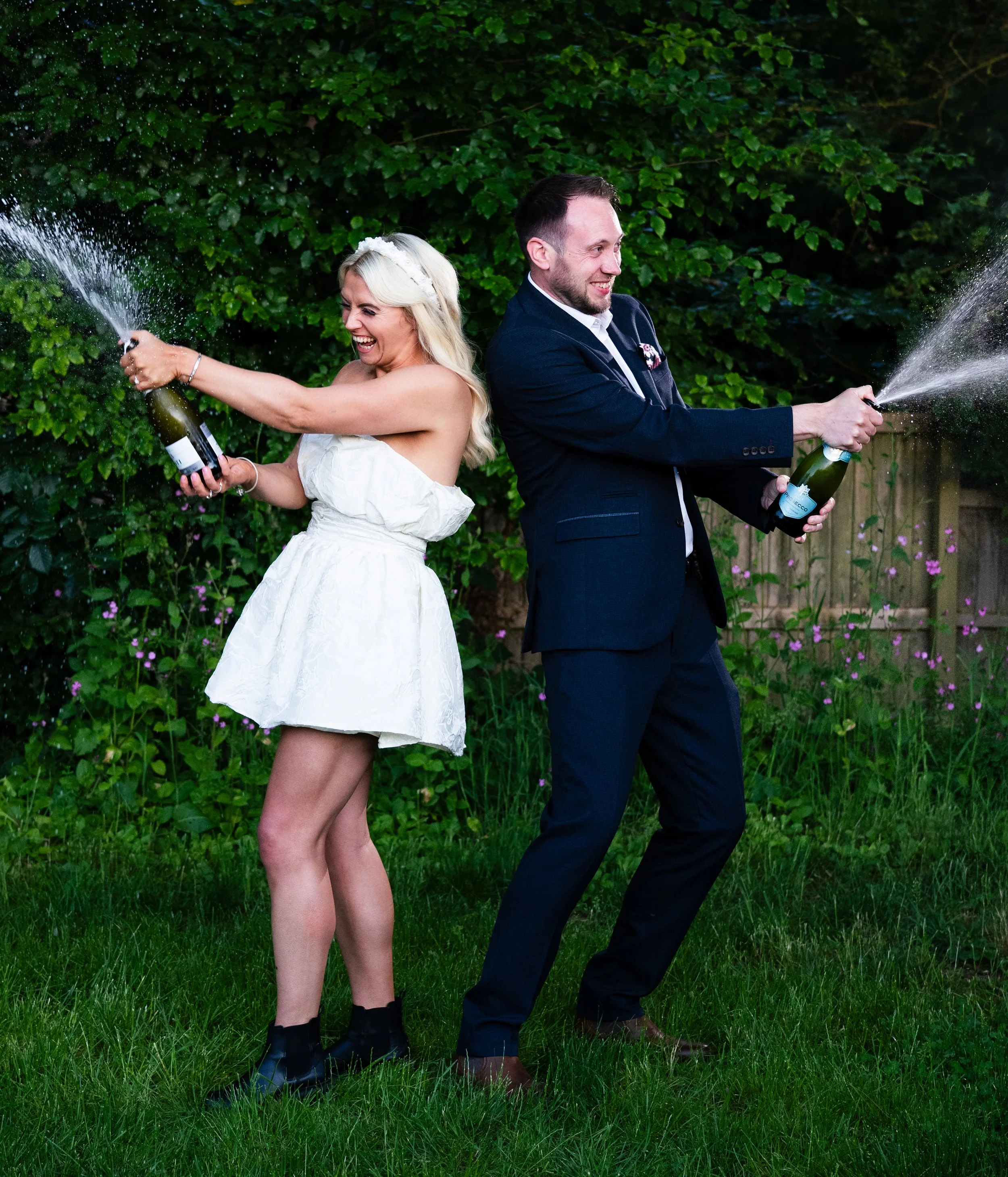 A bride and groom playfully spraying champagne at each other outdoors in a garden, holding a bottle of champagne with champagne sprays in the air, surrounded by green foliage and purple flowers.