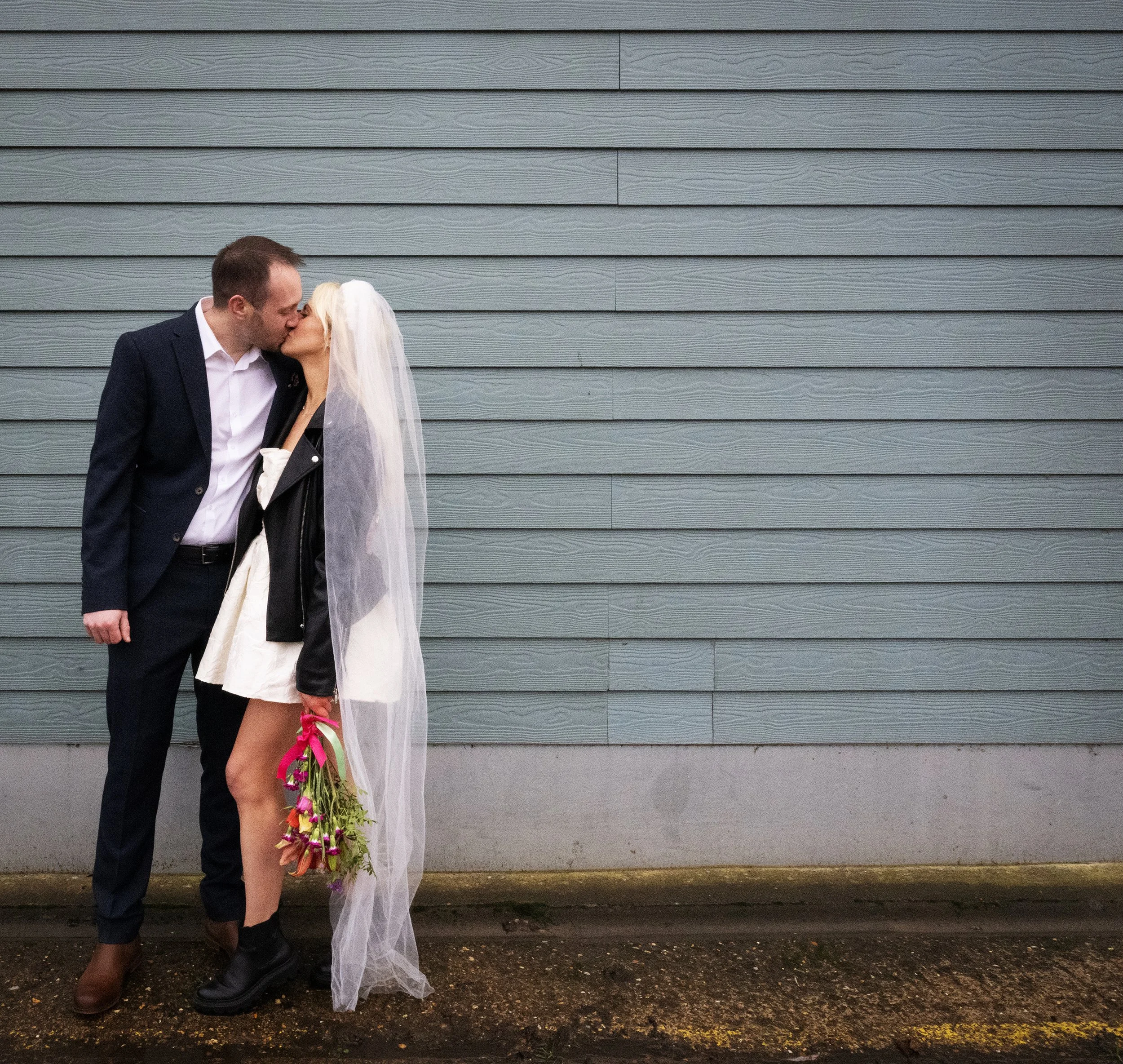 A bride and groom sharing a kiss in front of a blue wooden wall, the bride wearing a veil and holding a bouquet, and the groom dressed in a suit. Industrial alternative wedding 