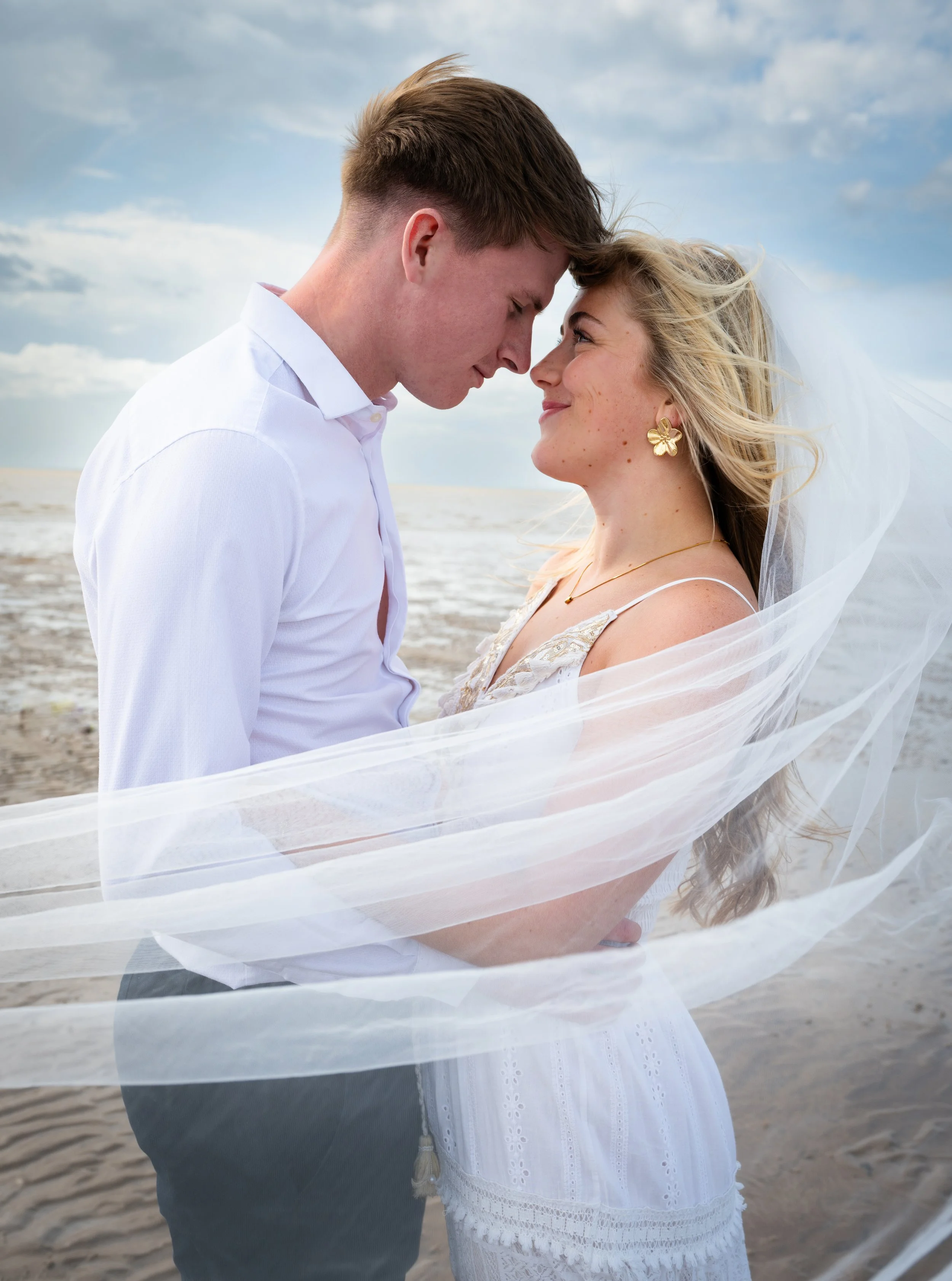 A couple on a beach, dressed in wedding attire, with their foreheads touching and smiling at each other, during daytime with a cloudy sky.