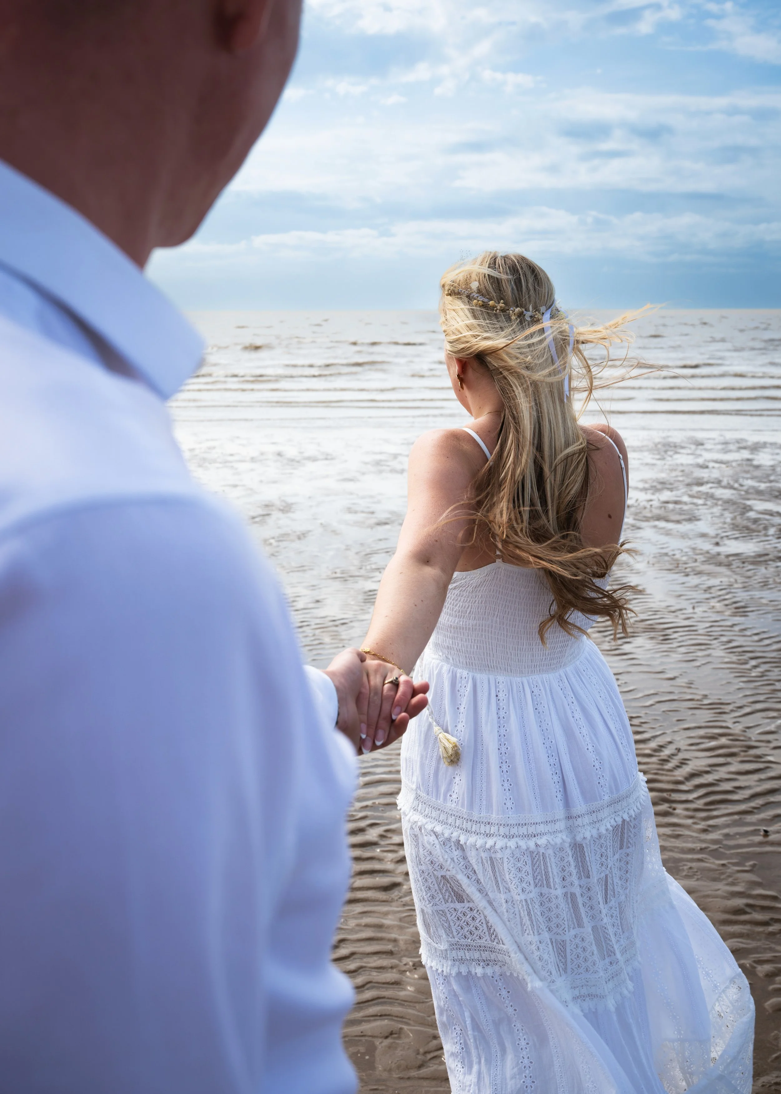 A couple holding hands on the beach, with the man in a white shirt and the woman in a white dress with lace details and a floral hair accessory, facing the ocean under a cloudy sky.