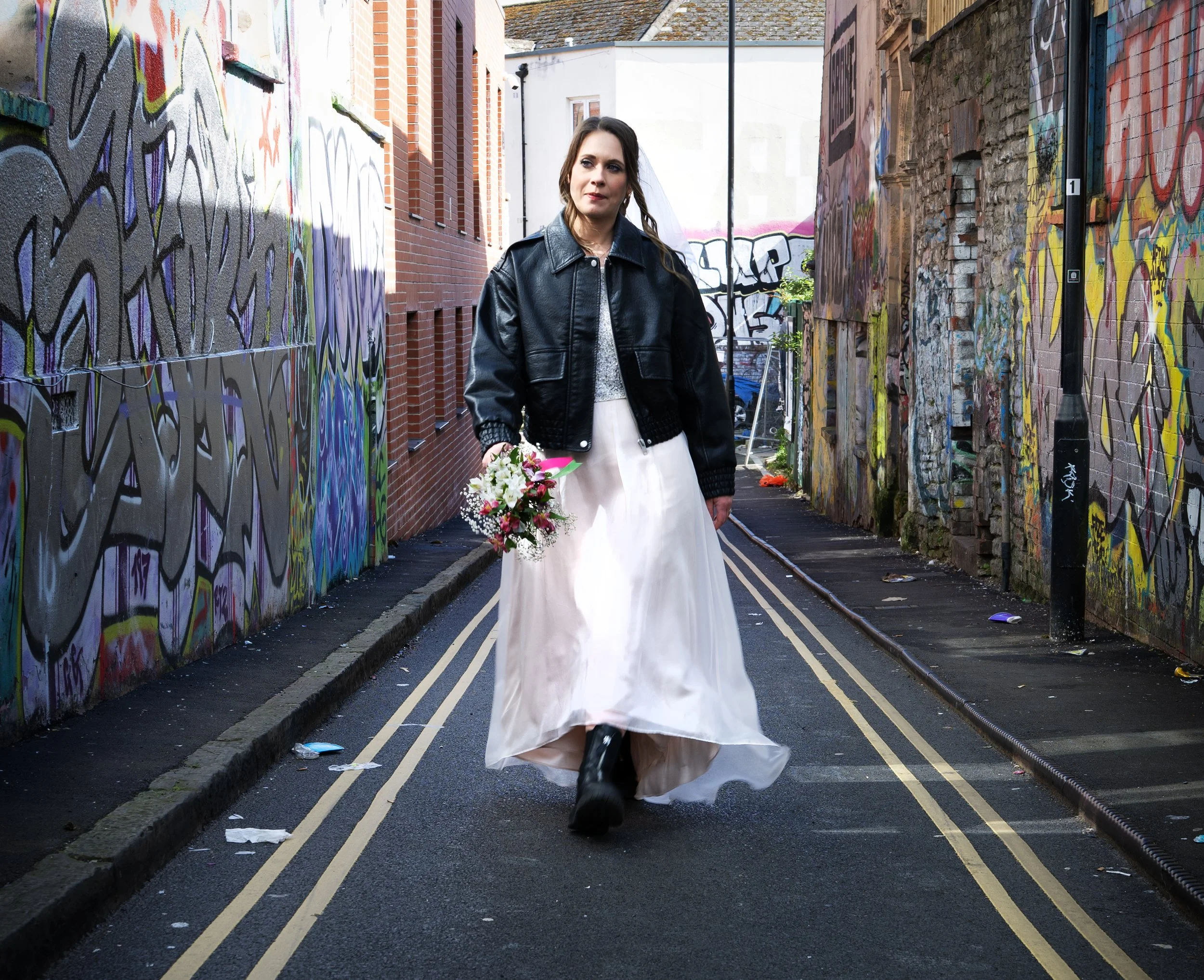 A bride wearing a white wedding dress, black leather jacket, and black boots walking down a graffiti-covered alleyway while holding a bouquet of flowers. Bristol Stokes Croft