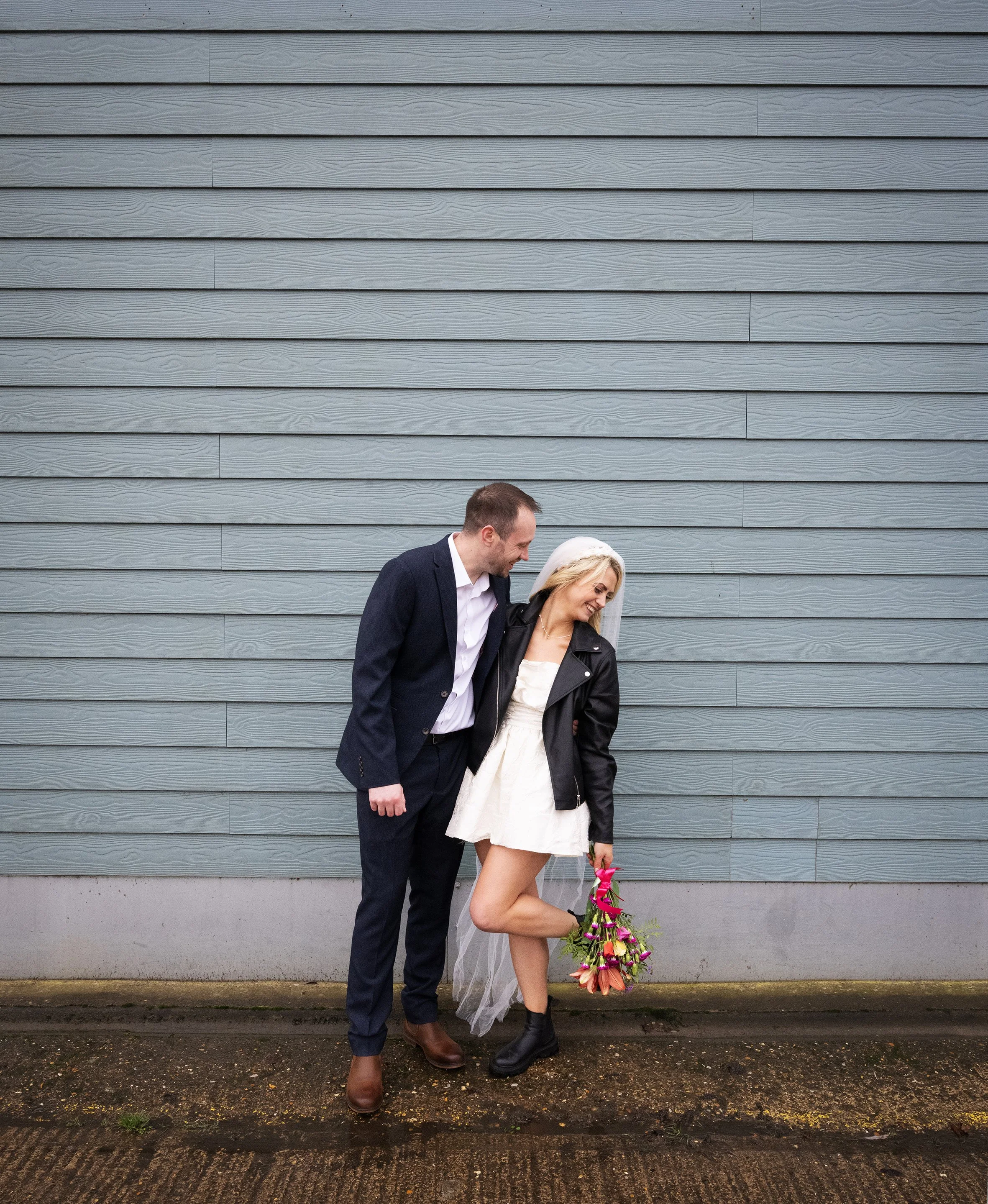 A couple standing on a wet pavement in front of a blue wooden wall, with the woman holding a bouquet of flowers. Bridal couple. Industrial wedding. Alternative Wedding