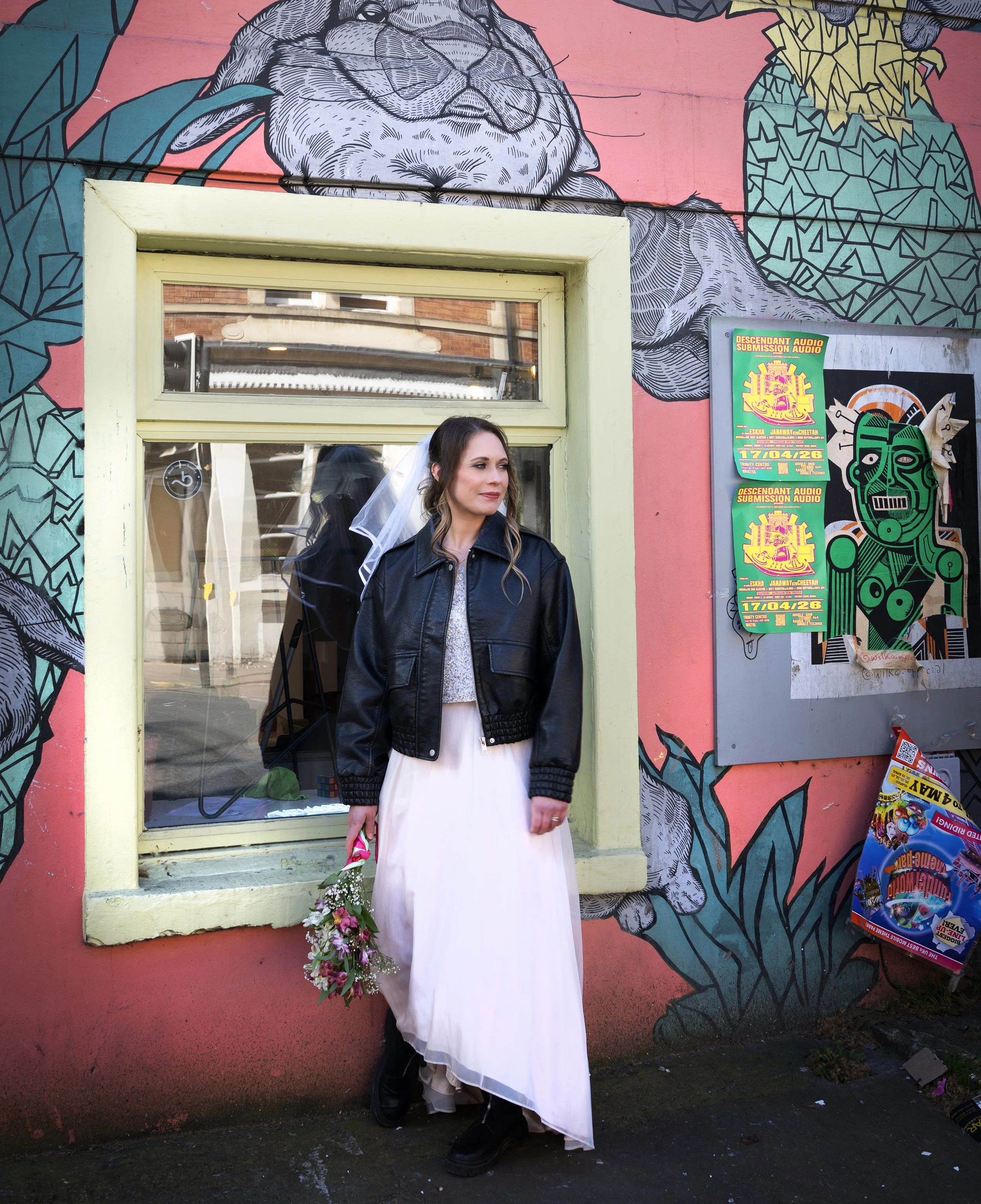 A bride in a white dress and black leather jacket holding a small bouquet of flowers, standing outside a colorful mural on a pink wall with artwork and posters, in front of a window. Bristol Stokes Croft