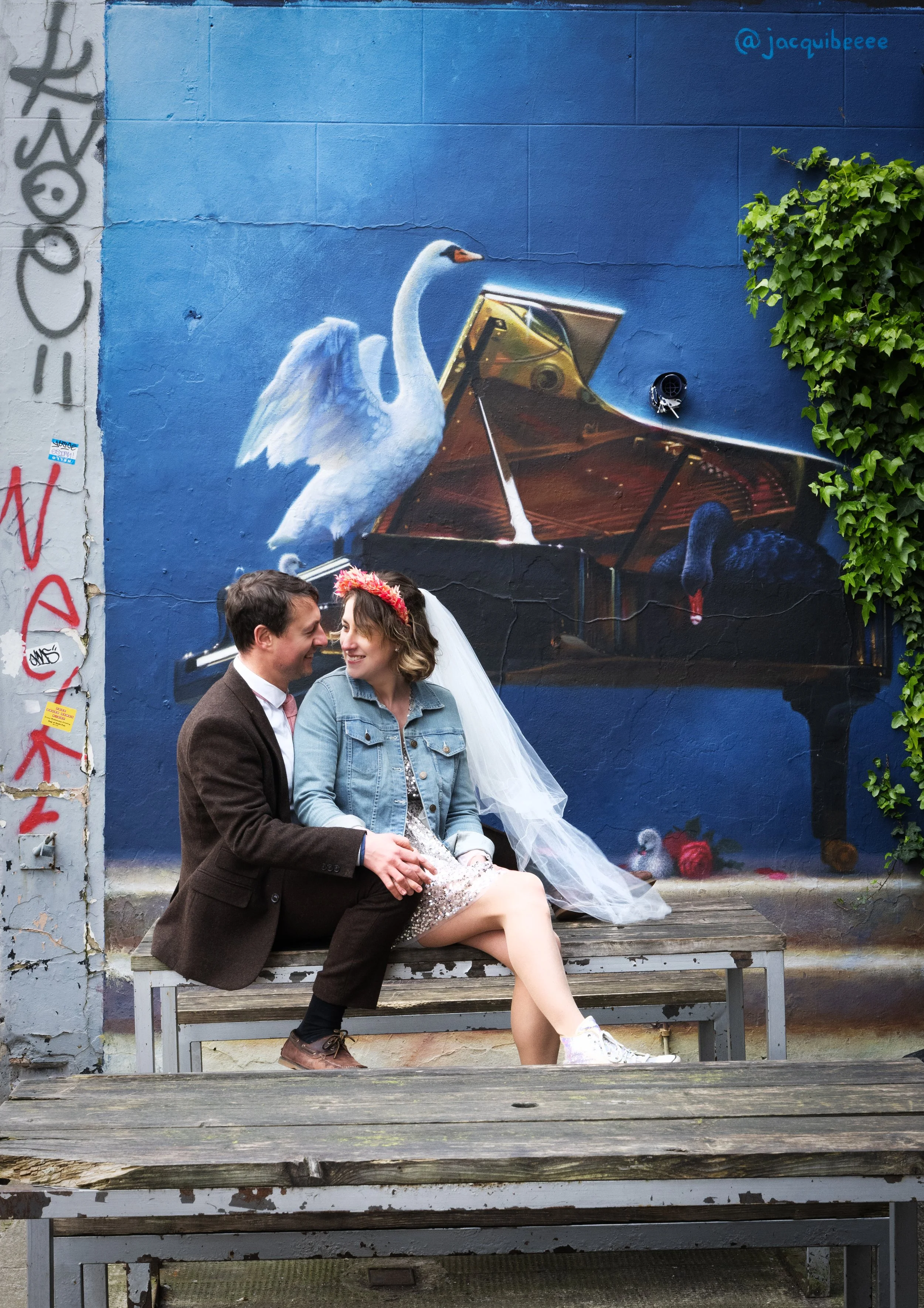 A wedding couple sitting on a bench with a mural of a swan, a black swan, and a grand piano painted on a blue wall behind them. Bristol North street