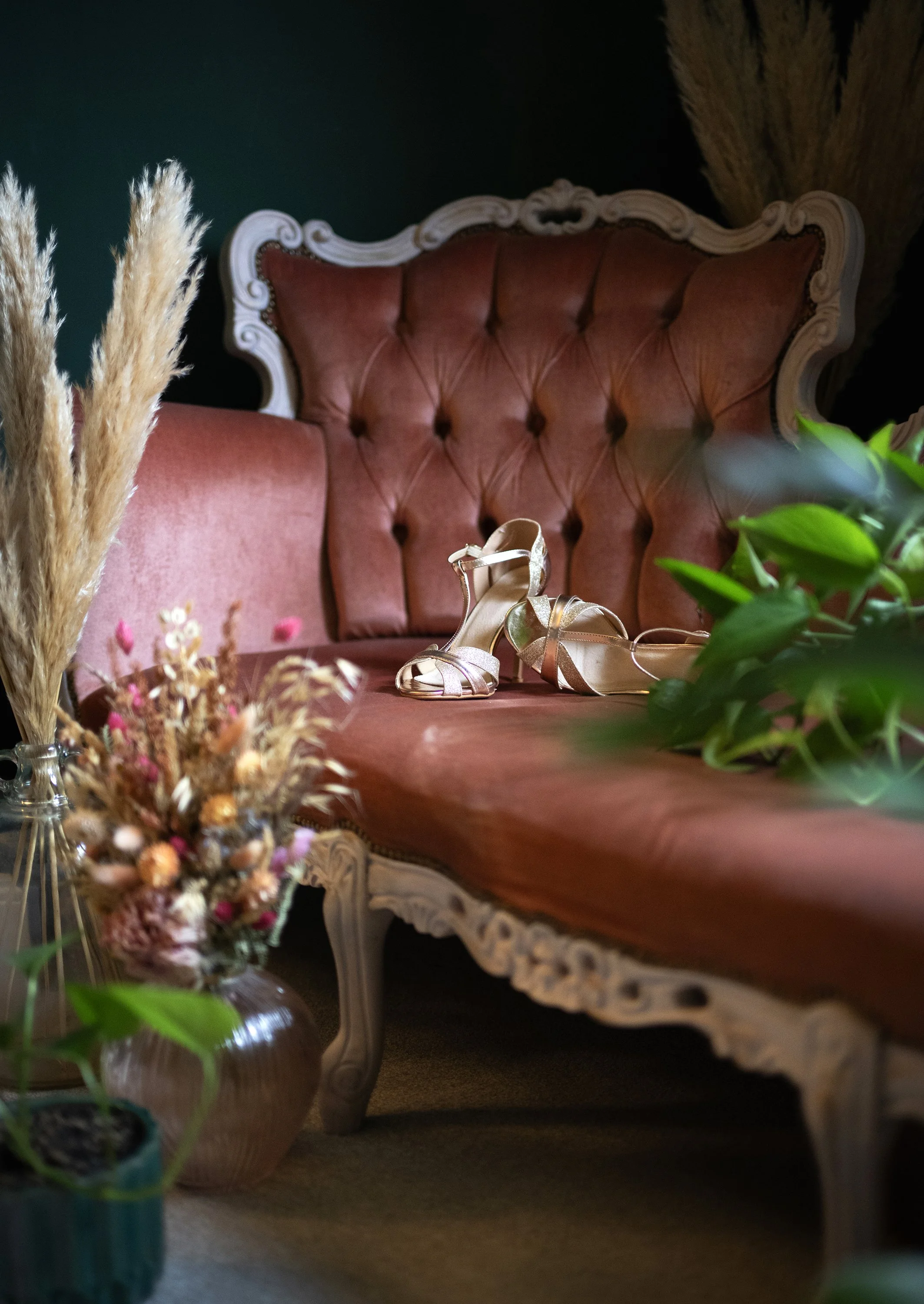 A vintage style pink velvet tufted sofa with ornate white wooden trim, a pair of gold high-heeled sandals resting on it, and surrounding plants and dried floral arrangements in vases. Vintage wedding. Alternative wedding. Timeless and Chic.