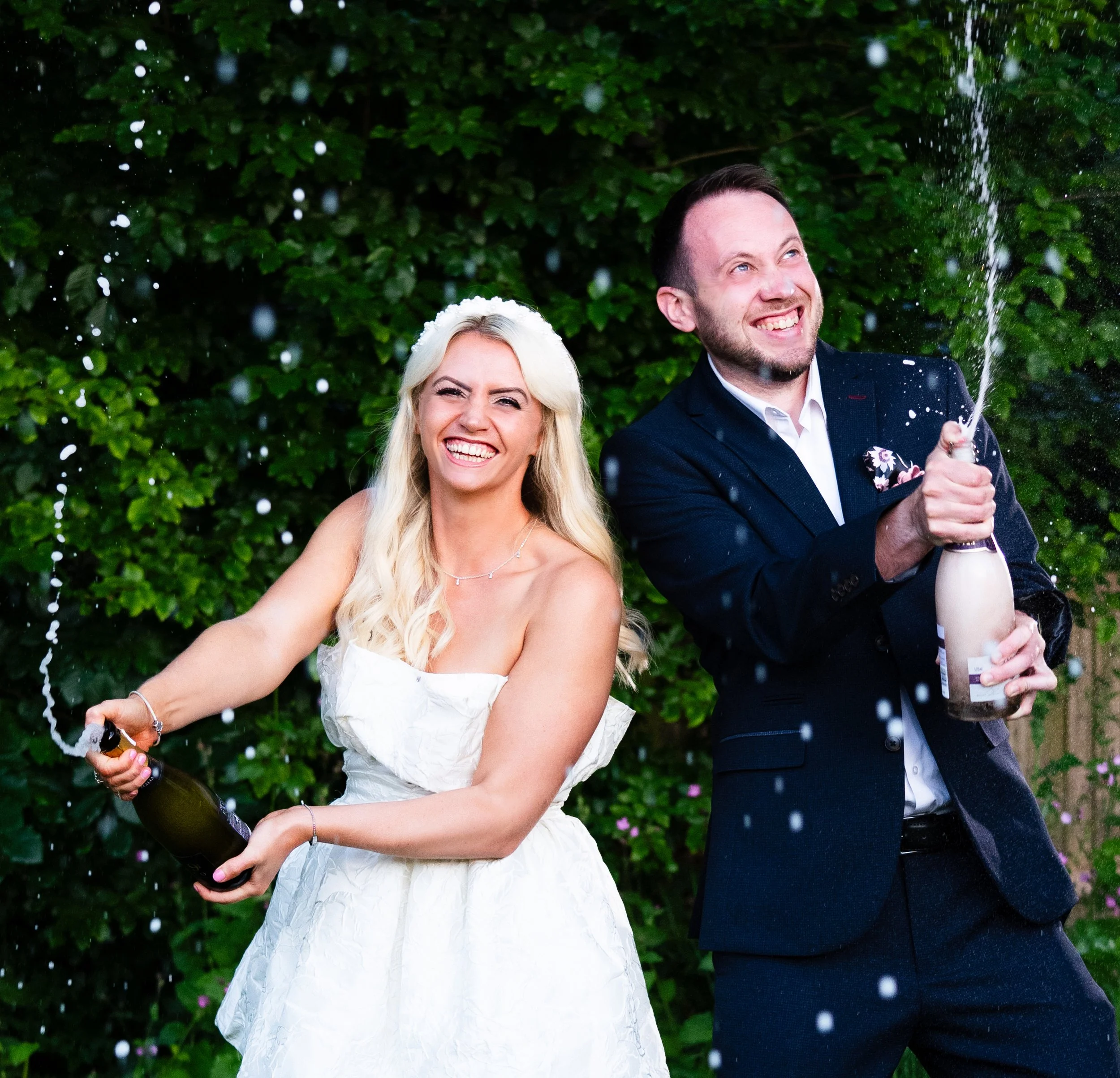 A joyful couple celebrating their wedding outside, with the groom opening a champagne bottle and champagne spraying, while the bride smiles happily in a white wedding dress, surrounded by green foliage.