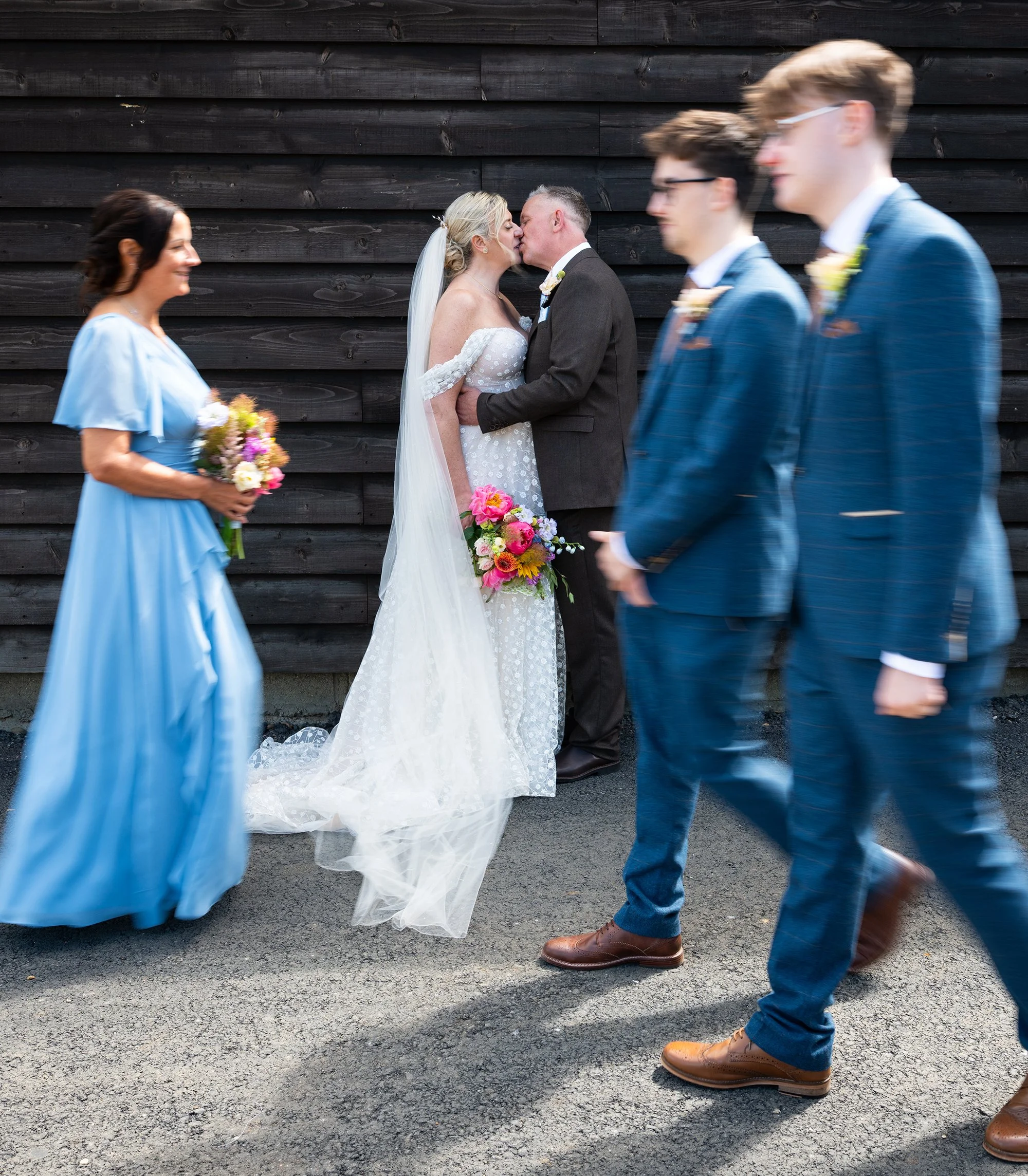 Bride and groom kissing while wedding guests walk past in the foreground.