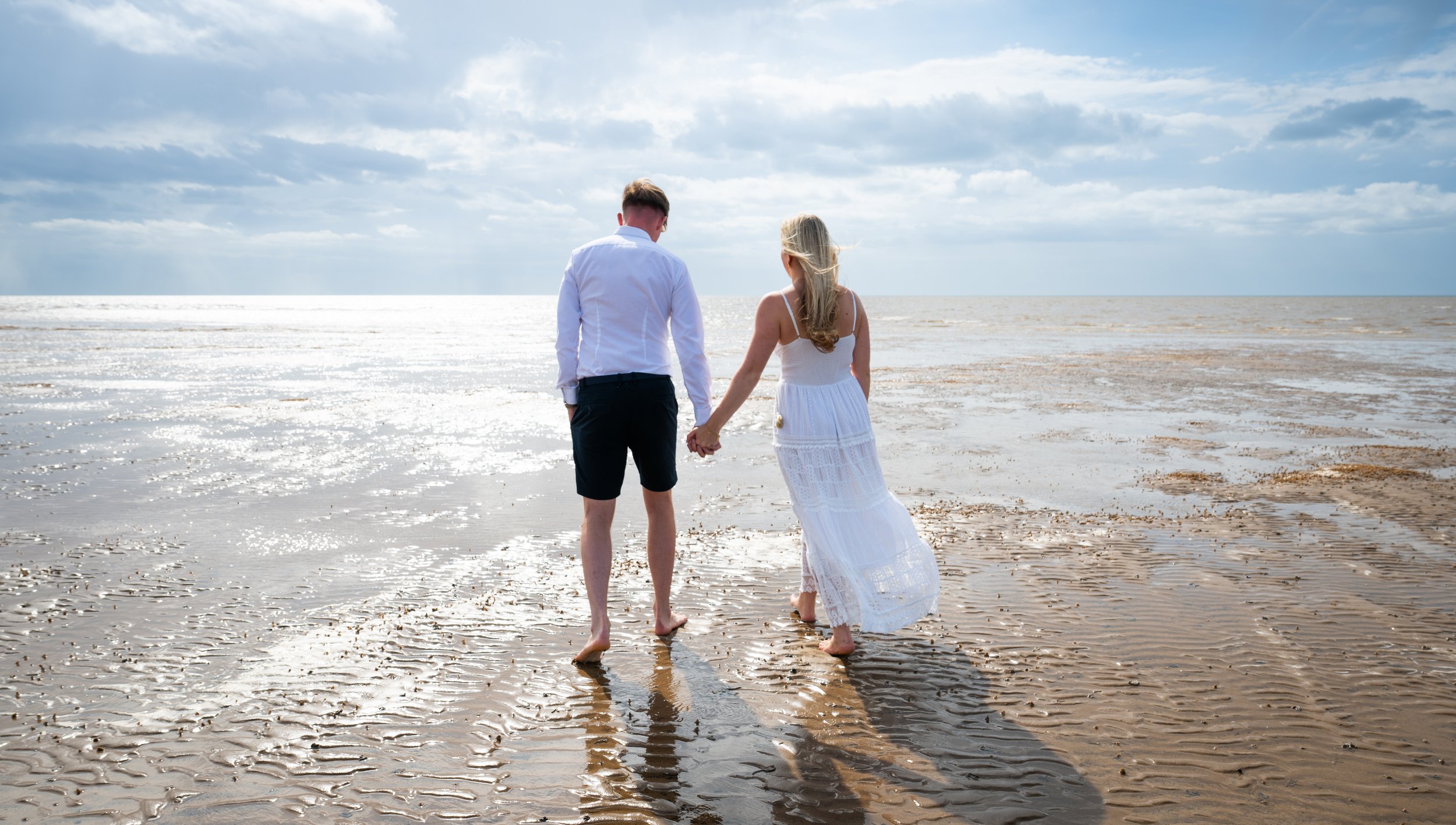 A couple, a man and a woman, holding hands and walking barefoot on a sandy beach near the ocean, with clouds in the sky.