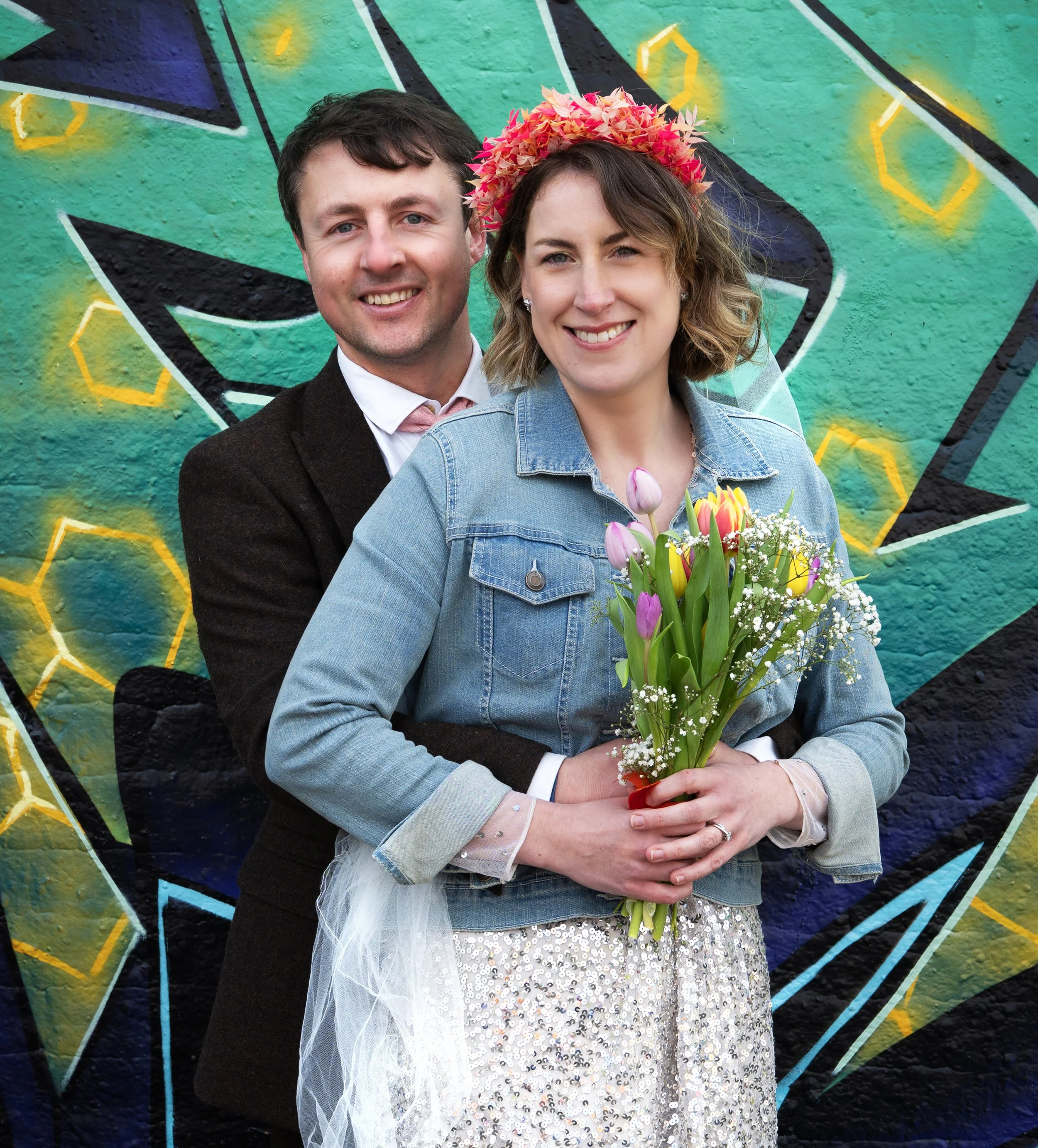 A smiling couple stands in front of a colorful graffiti wall, with the woman holding a bouquet of tulips and wearing a floral crown. Dean Lane Skatepark Bristol Wedding