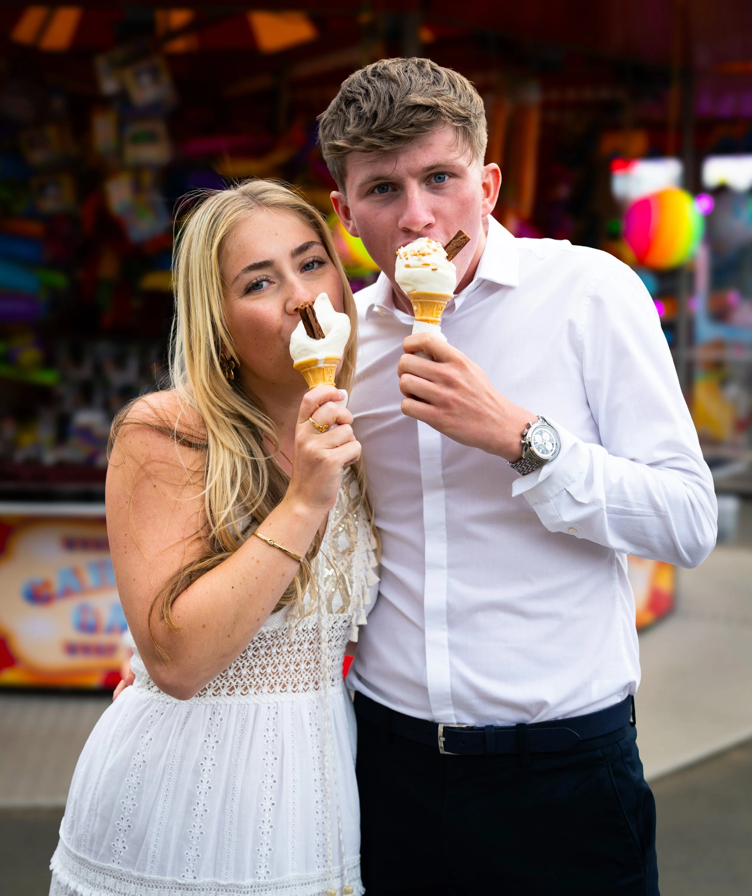 A young woman and a young man are standing close together, each holding an ice cream cone. The woman has blonde hair and is wearing a white, eyelet dress. The man has short, curly hair, and is dressed in a white shirt and dark trousers, with a silver