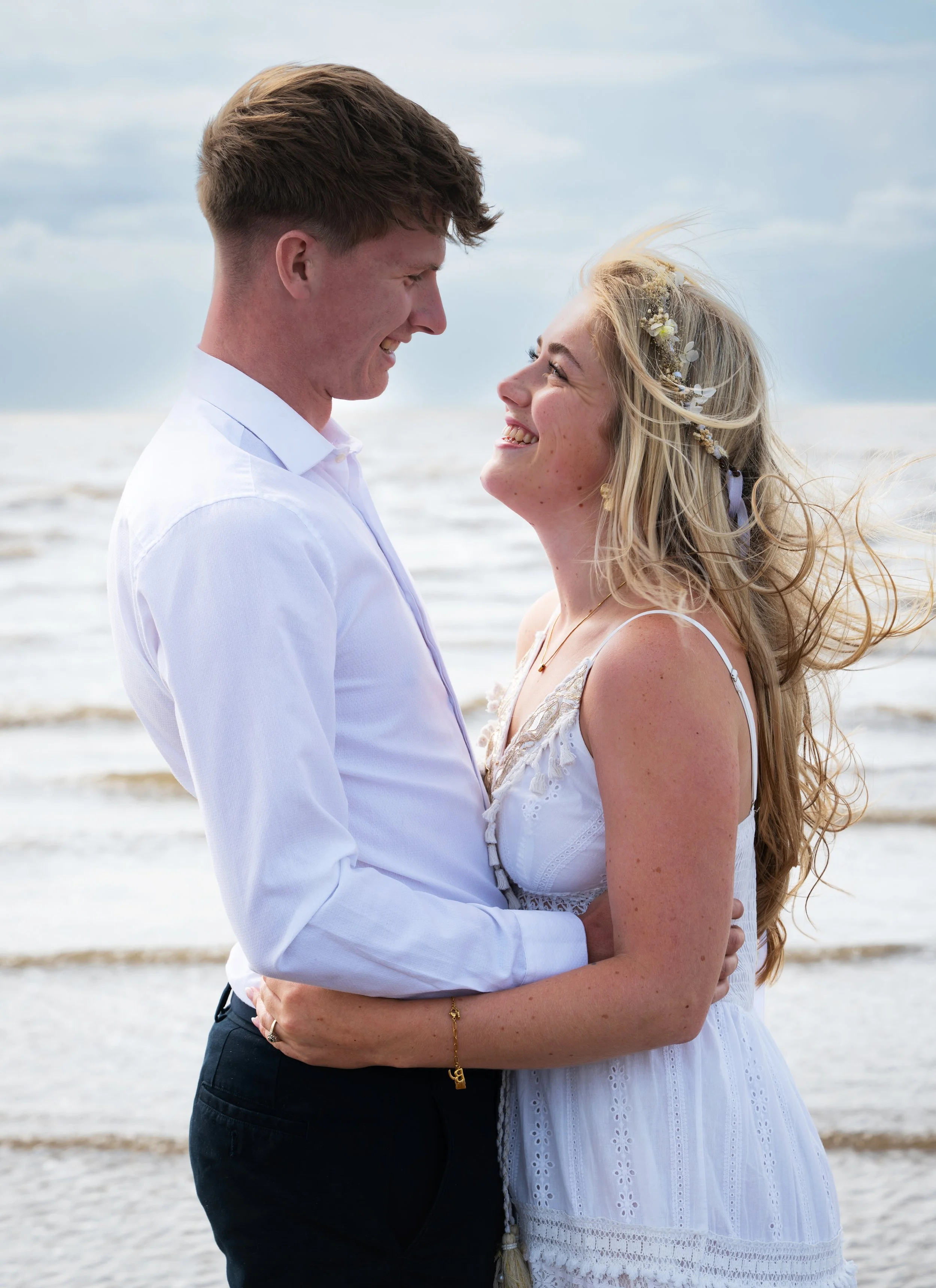 A young couple at the beach, smiling and embracing each other.