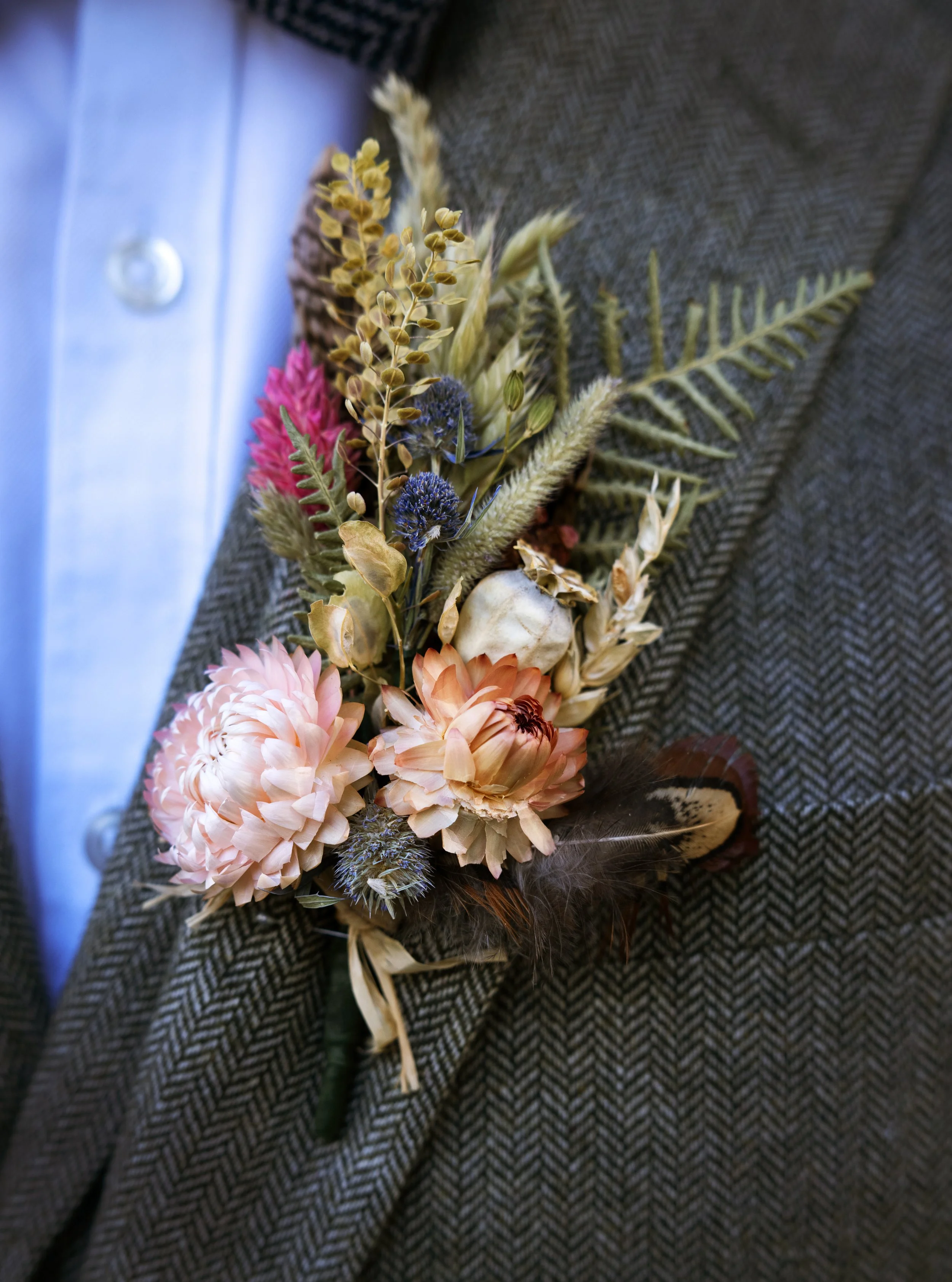 Close-up of a boutonniere with pink and beige flowers, dried grass, fern leaves, wrapped with a brown feather, pinned to a brown tweed jacket, with an light blue shirt underneath. Groom buttonhole. Groom accessories. Groom detail shots.