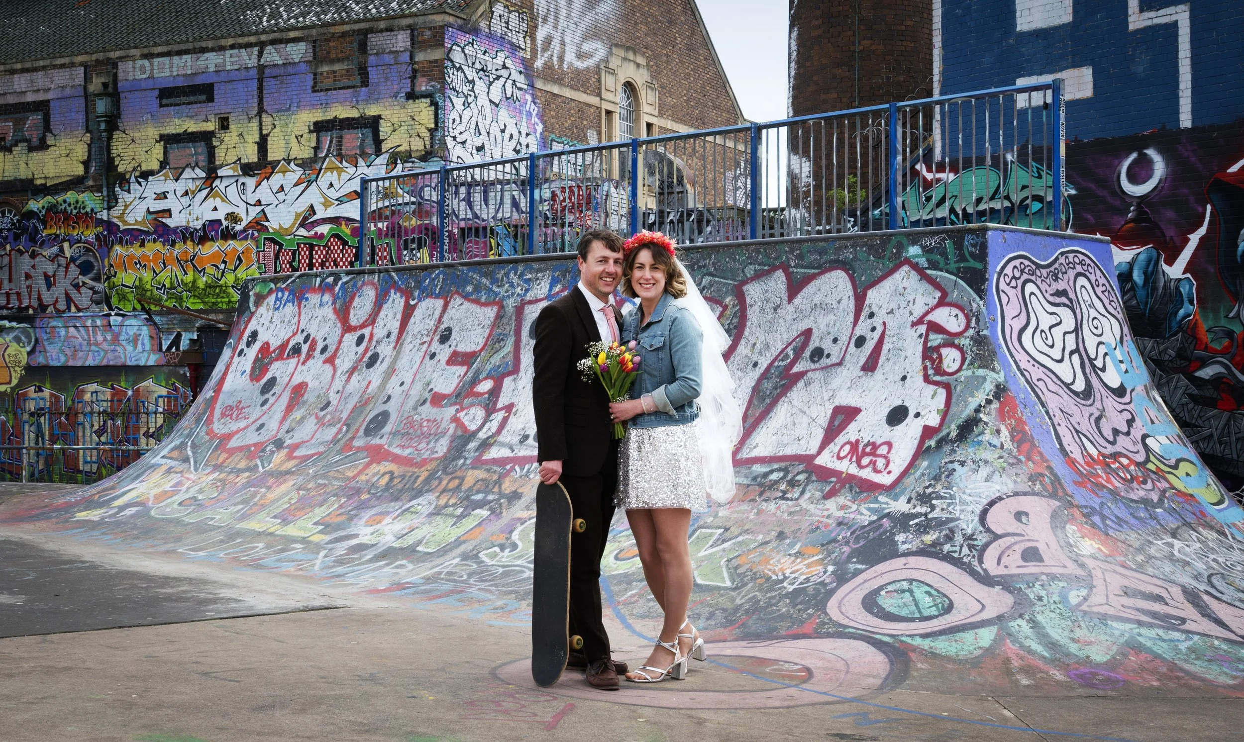 A newlywed couple standing side by side at a graffiti-covered skate park. The groom is holding a skateboard, and the bride is holding a bouquet of colorful flowers. They are smiling, with the groom in a black suit and the bride in a white sequined dr