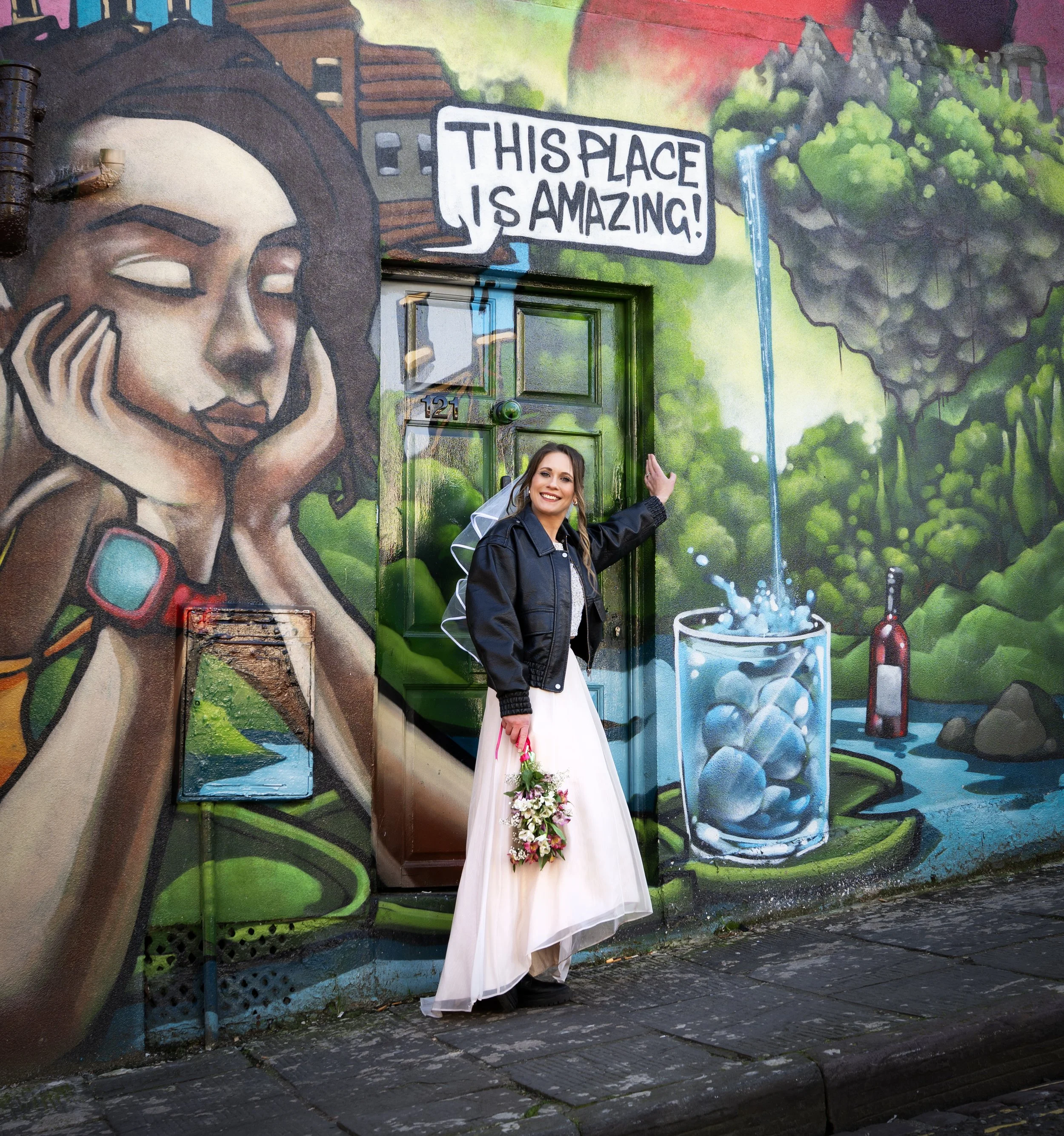 A bride in a white dress and black jacket holding a bouquet of flowers, smiling and pointing at a mural on a wall behind her. Bristol Stokes Croft