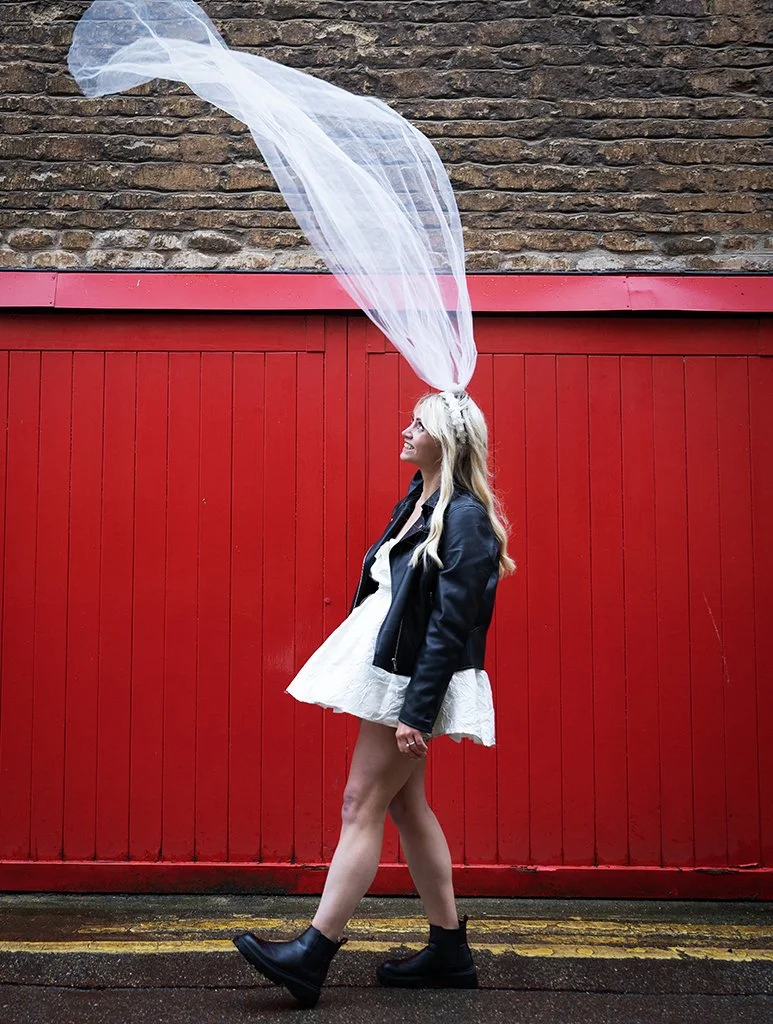Alternative bride walking past a bright red wall with her veil blowing in the wind.