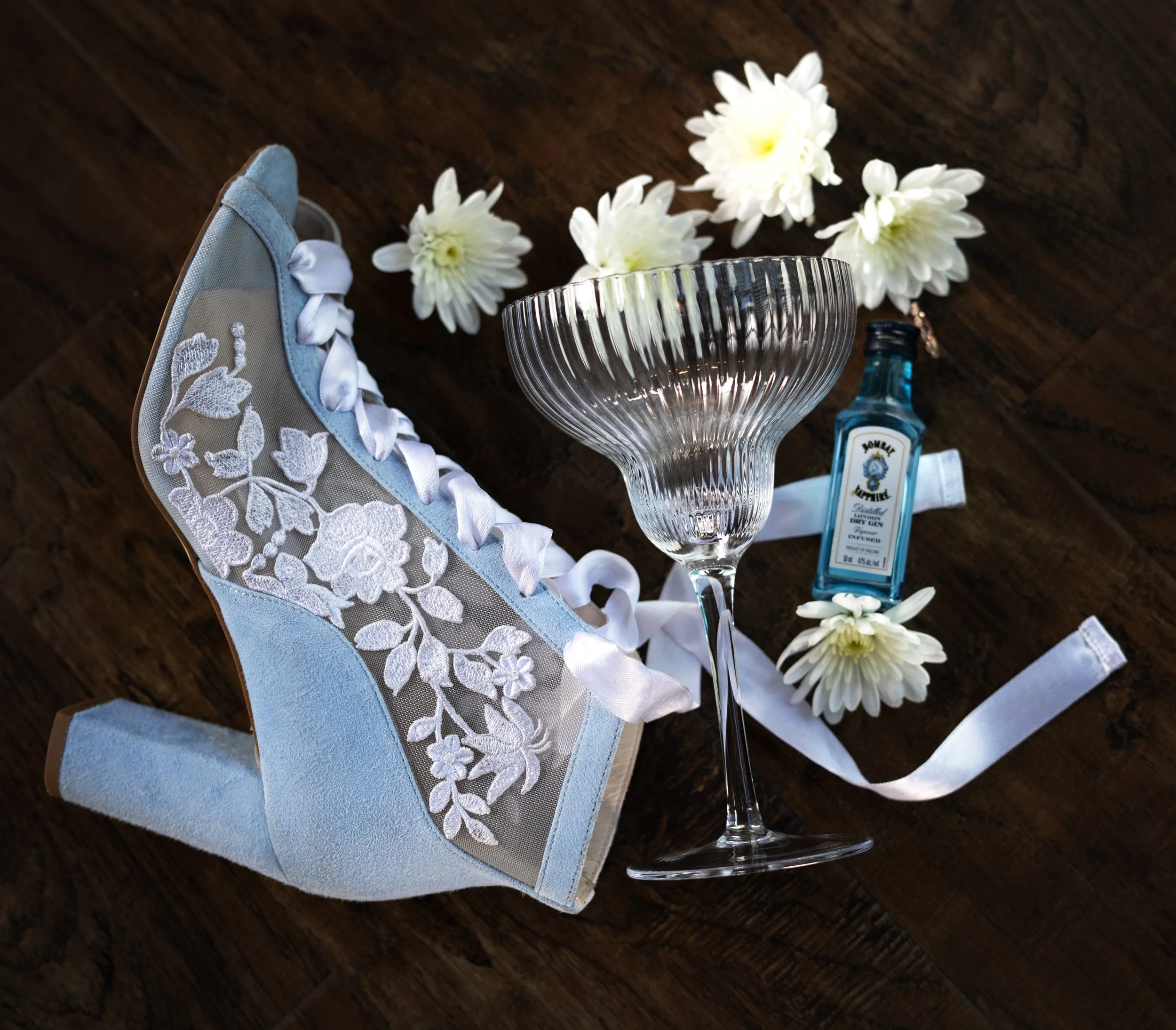 A delicate blue high heel shoe with white floral embroidery, white ribbons, a clear cocktail glass, white flowers, a blue bottle of Bombay Sapphire gin, and white ribbons on a dark wooden surface. Bridal details. Wedding accessories. Wedding flatlays