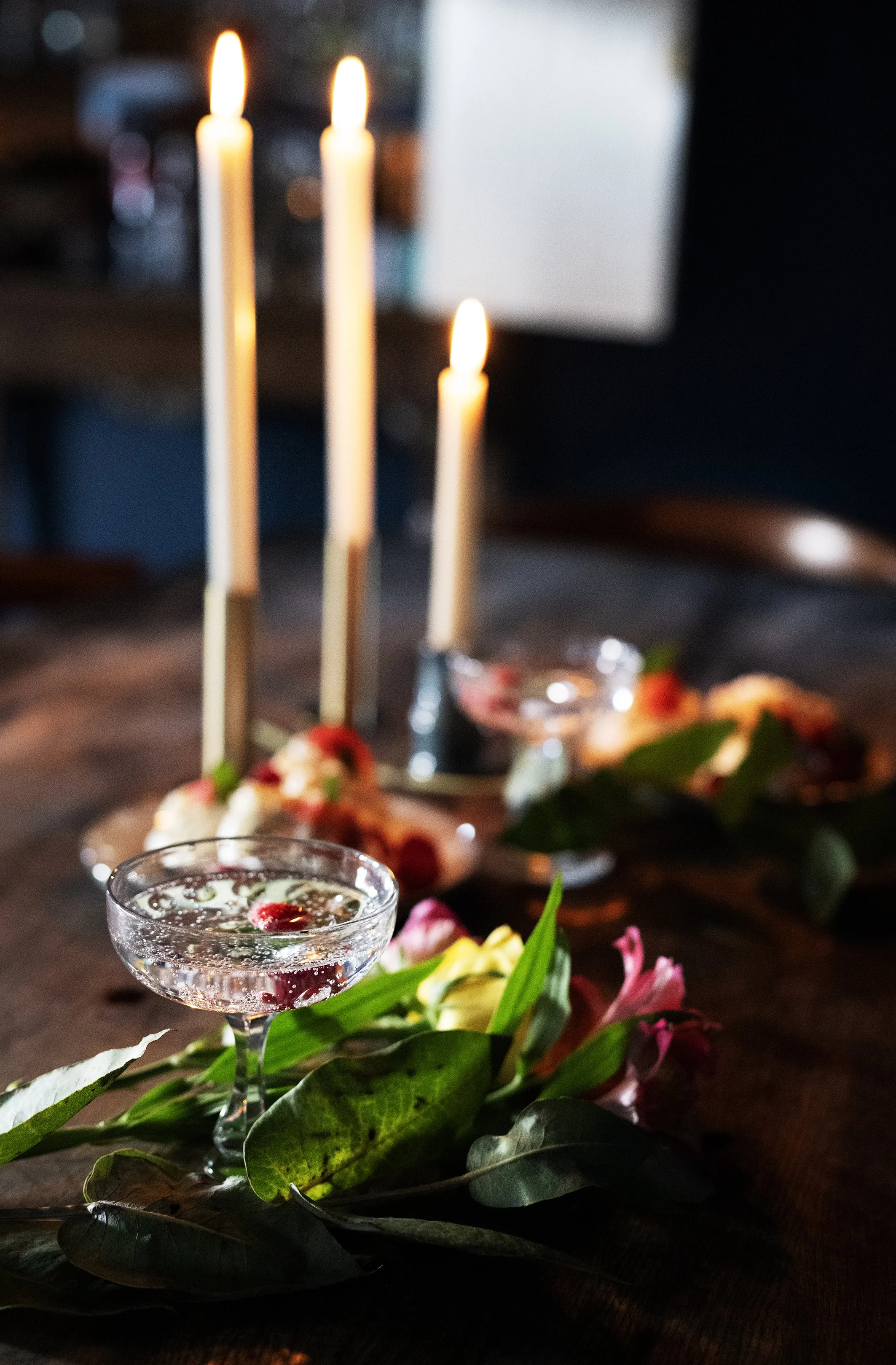 A table with three lit candles, a champagne coupe glass filled with a bubbly drink, and floral decorations. Wedding table details. Wedding breakfast.