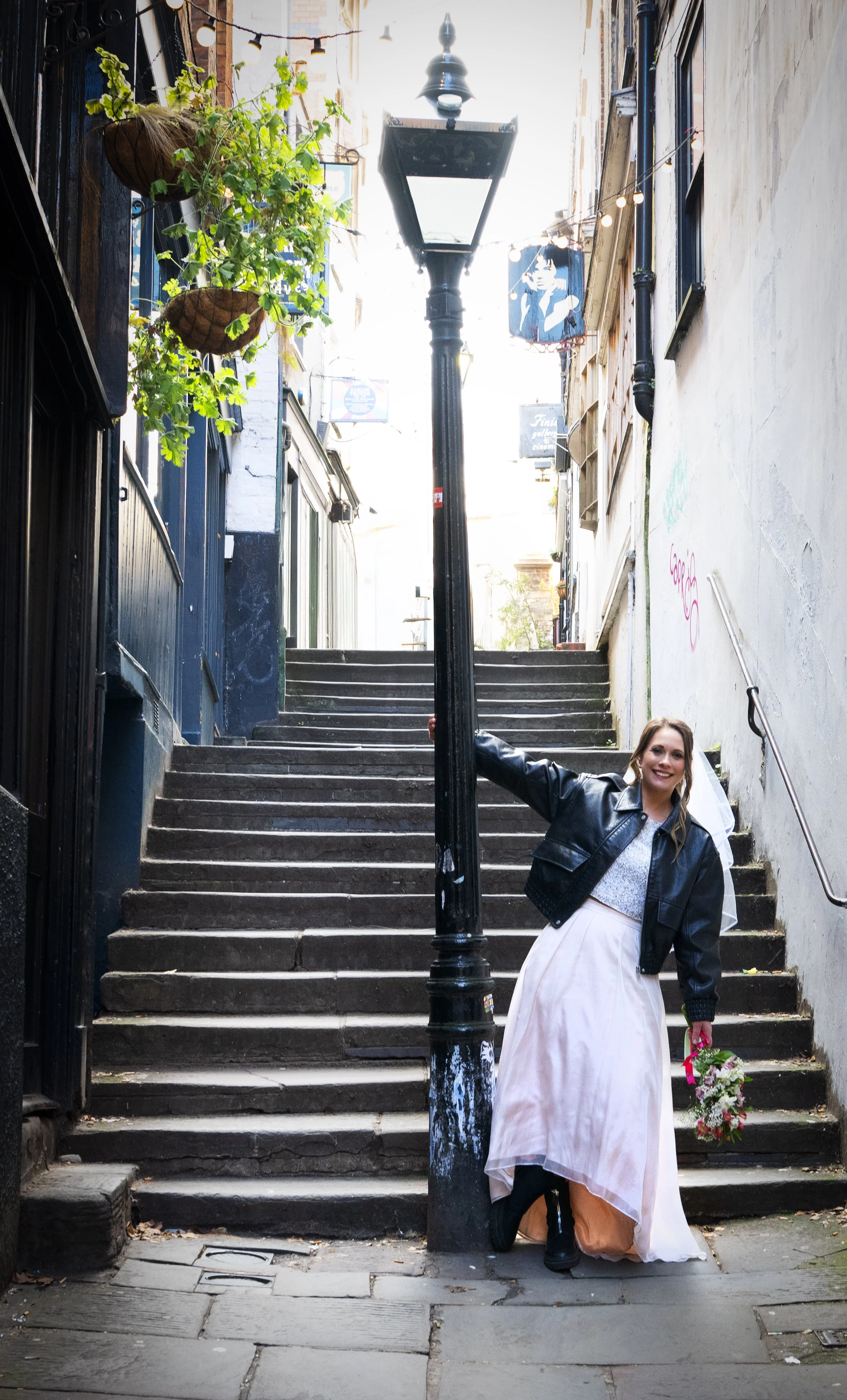 A bride in a wedding dress and leather jacket holding a bouquet, leaning on a street lamp on a city back alley staircase, smiling. Bristol Christmas Steps