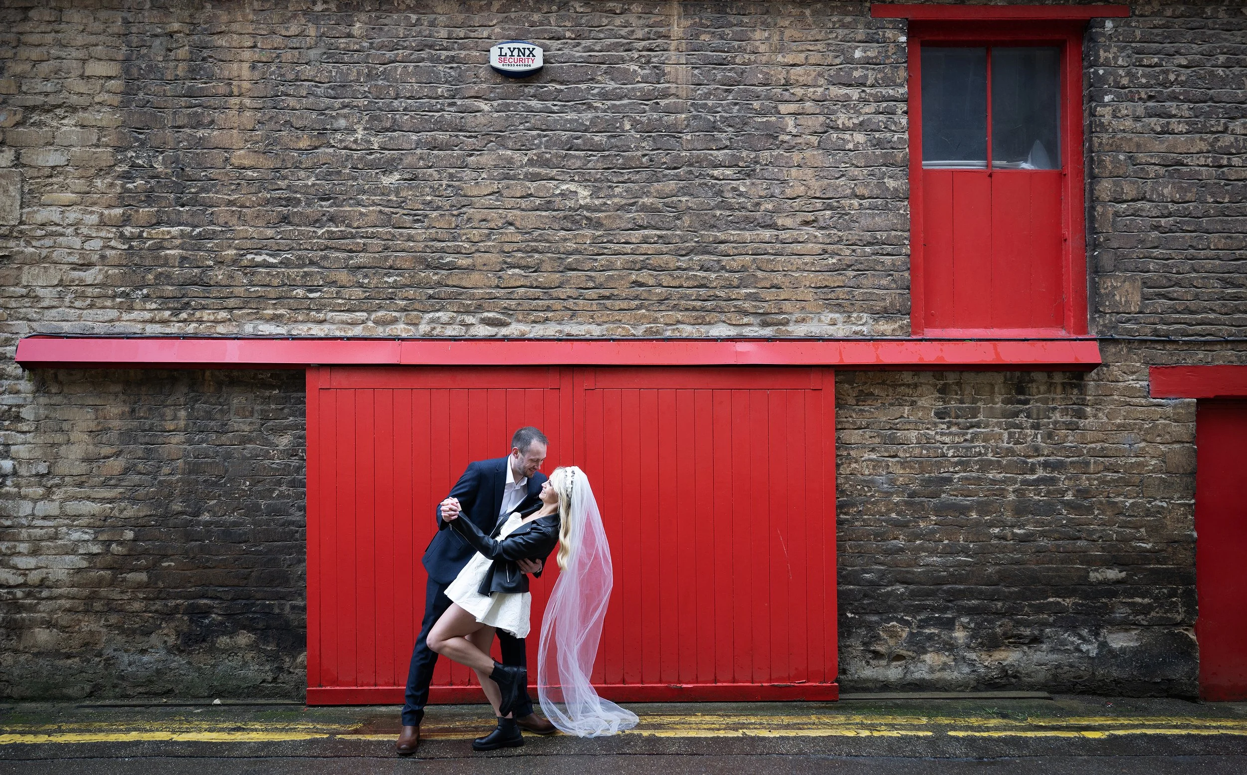A bridal couple dancing romantically in front of a red garage door and brick wall, with the bride wearing a veil and black boots, and the groom in a black suit. Industrial wedding. Alternative wedding.