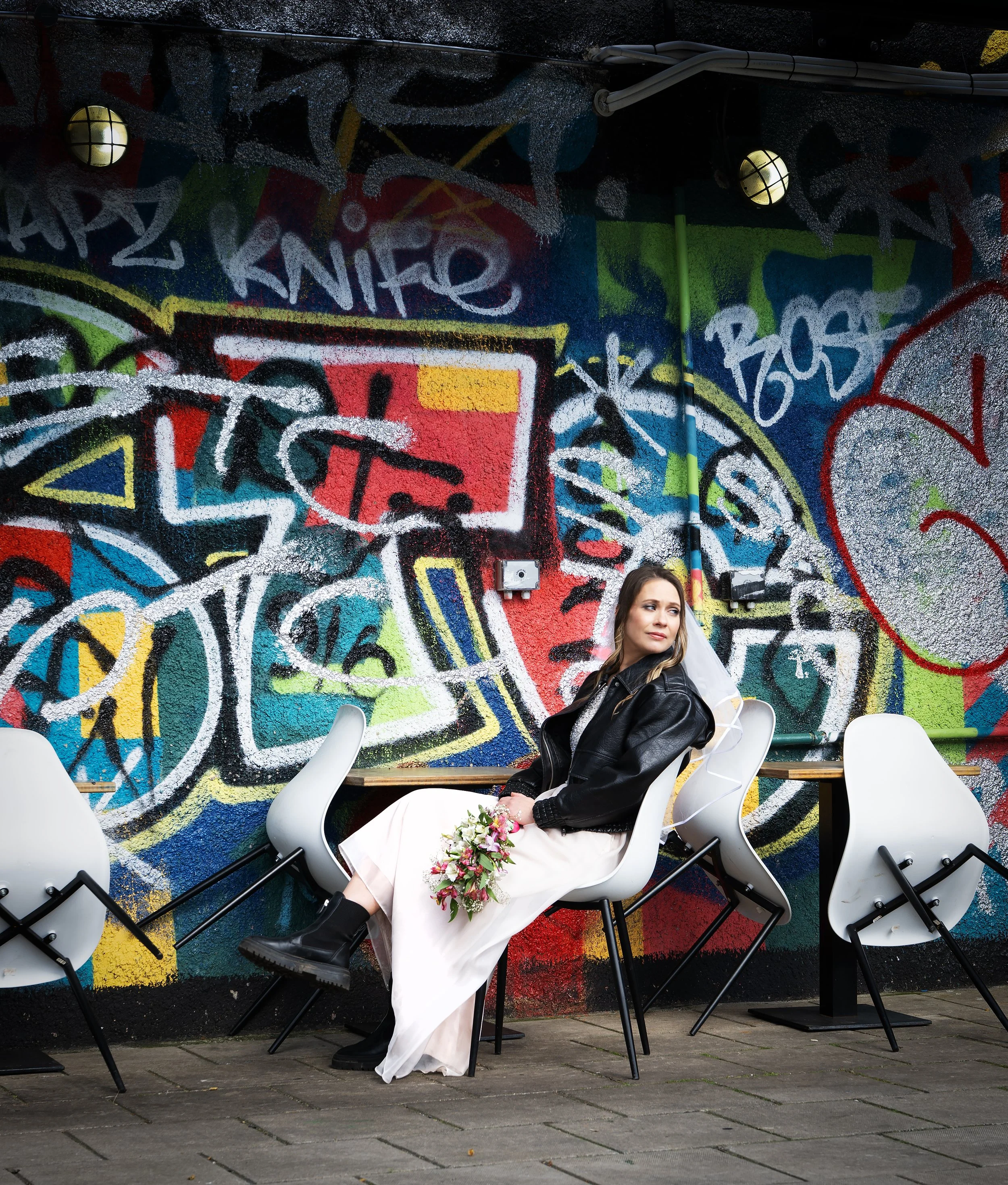 A bride in a white dress, black leather jacket, and black boots sitting on a modern white chair, holding a bouquet of flowers, outside against a vibrant graffiti wall. Bristol Stokes Croft
