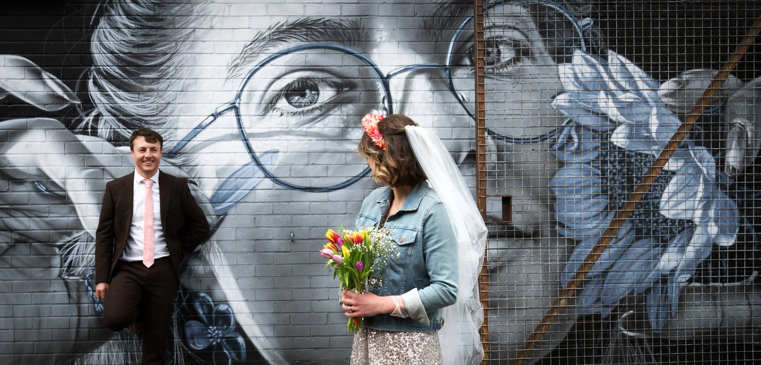 A bride holding a bouquet of tulips and wearing a veil and flower headband, looking at the groom in a suit standing against a graffiti wall featuring a large portrait of a woman with glasses. Bristol North Street