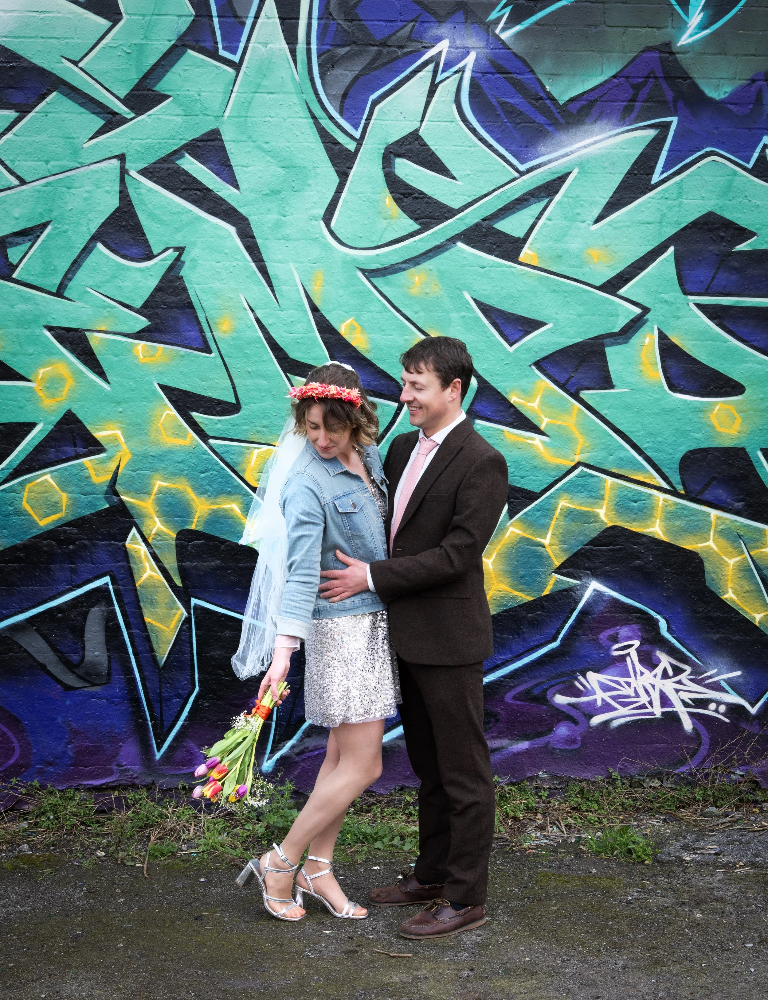 A couple standing in front of a colorful graffiti wall, with the woman holding a bouquet of tulips, smiling and looking down as the man holds her around the waist, both dressed in stylish outfits. Dean Lane Skatepark Bristol