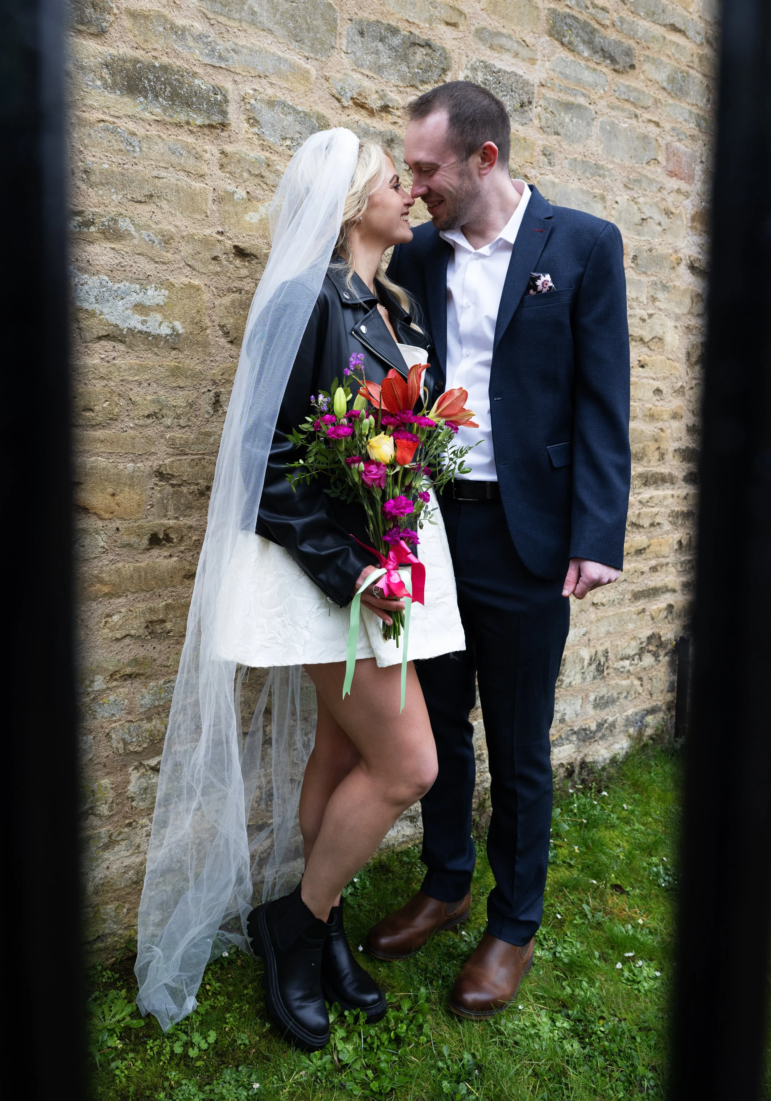 A couple stands close together against a brick wall, smiling and touching foreheads. The woman, wearing a white dress, black combat boots, a leather jacket, and a wedding veil, holds a colorful bouquet of flowers with pink, orange, and yellow blooms.