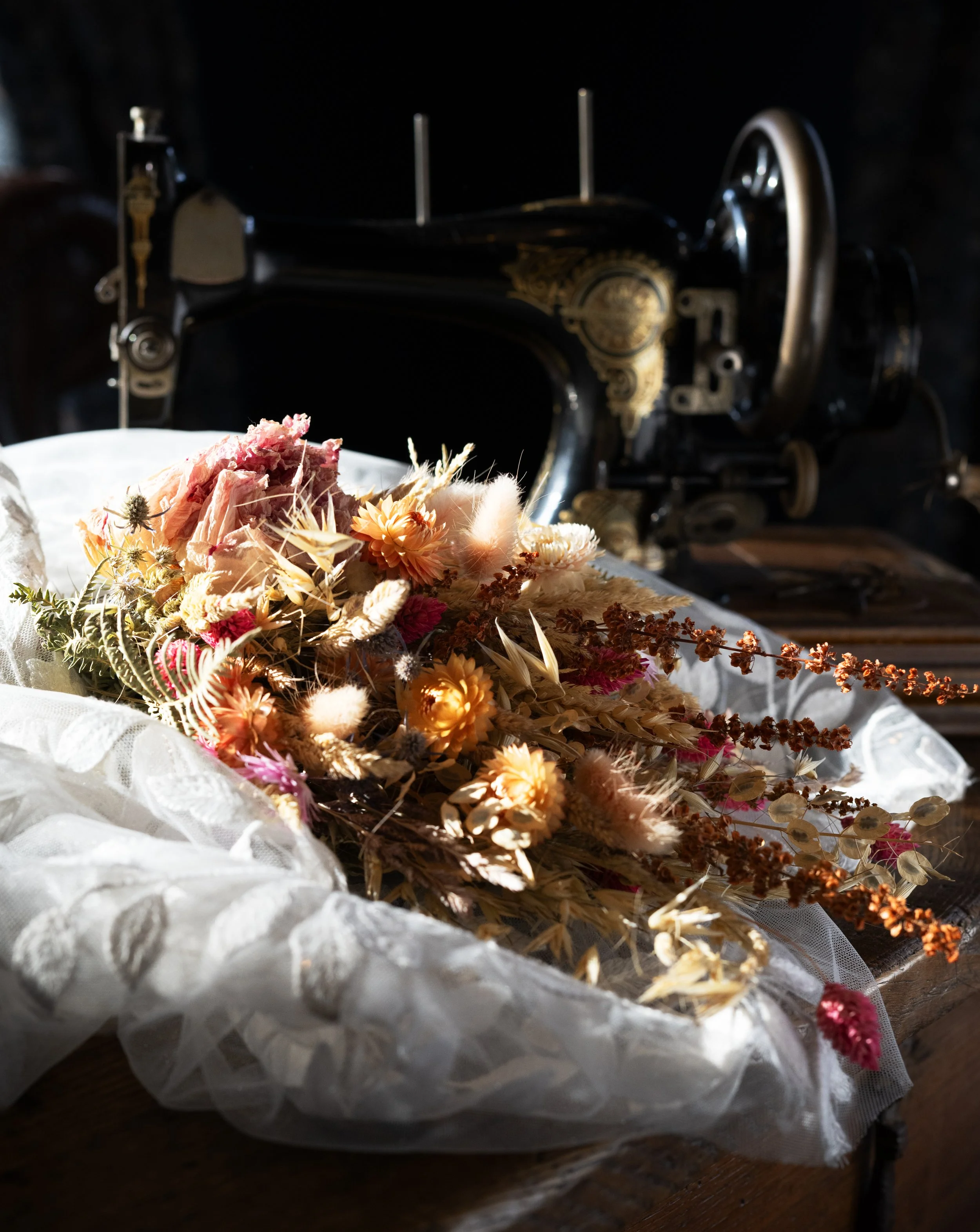 A vintage black sewing machine with gold detailing, embroidery, and a large wheel, with a bouquet of dried flowers in soft pastel and earth tones on a wooden surface illuminated by natural light. Vintage wedding. Timeless chic. Wedding bouquet.