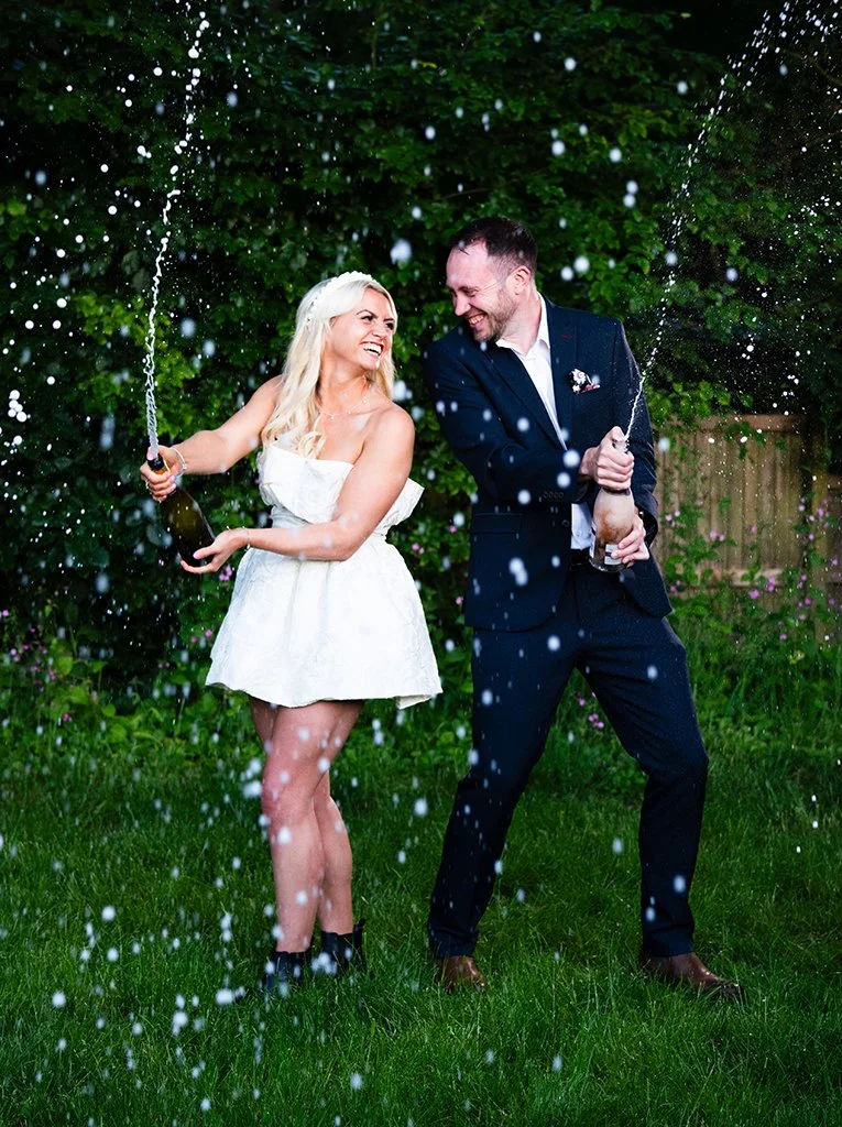 Bride and groom laughing while spraying champagne during an outdoor wedding celebration.