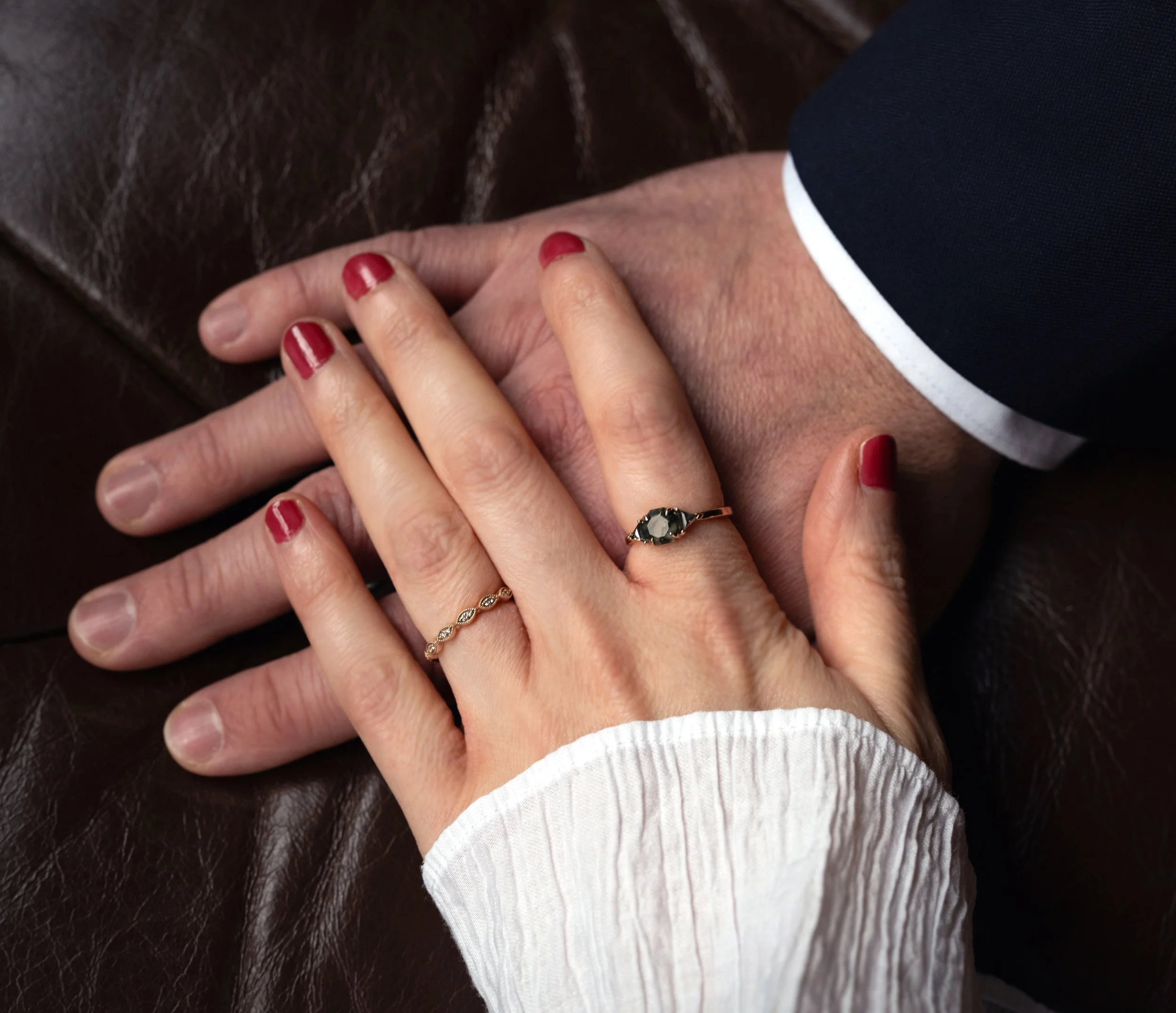 Close-up of wedding couples hands with red nail polish, wearing rings, resting on a brown leather surface with part of a person's arm in a dark suit visible.