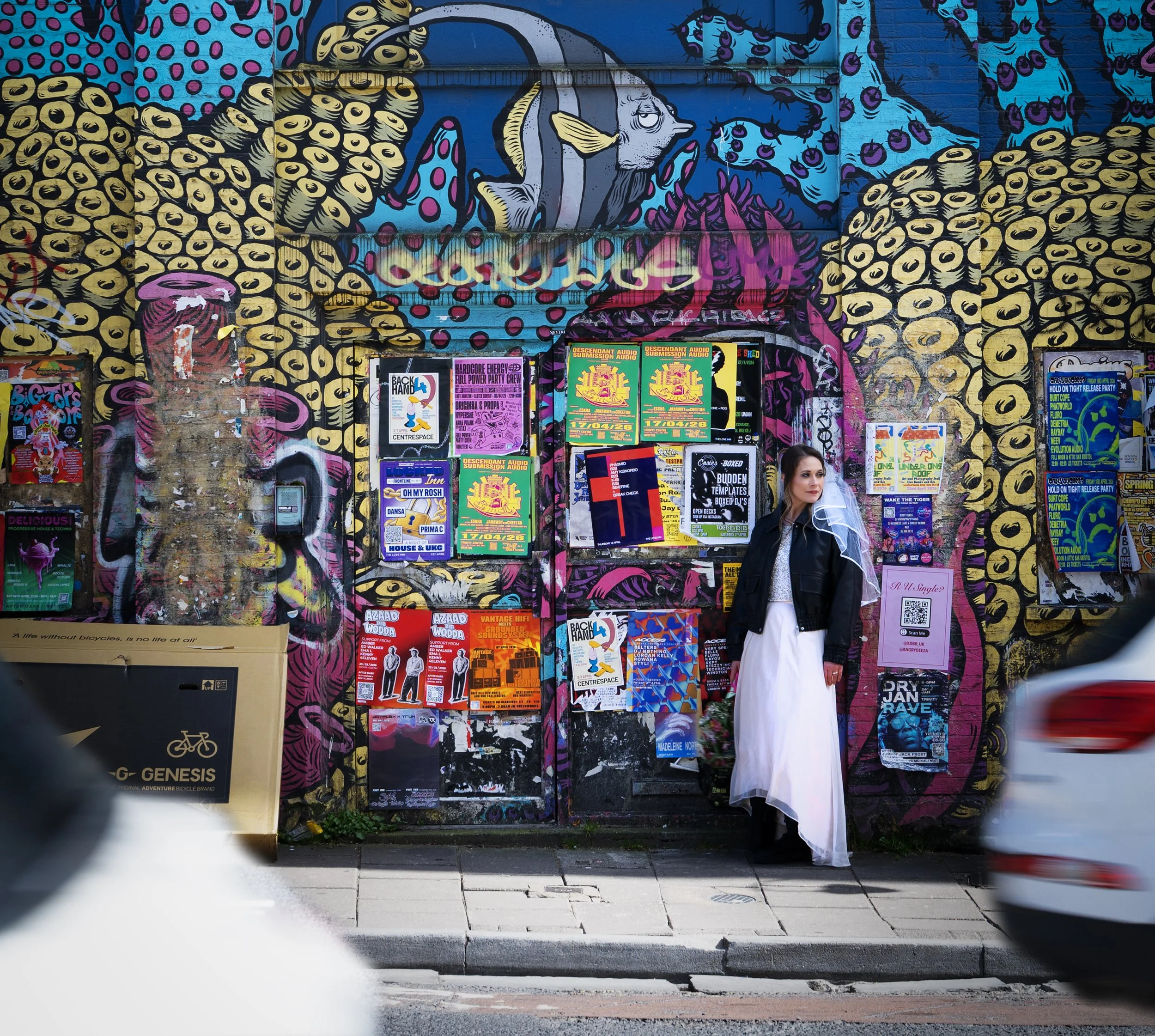 A bride in a white dress and black leather jacket standing against a vibrant, graffiti-covered wall with various posters and advertisements, part of which features a colorful, abstract mural of a fish. Bristol Stokes Croft