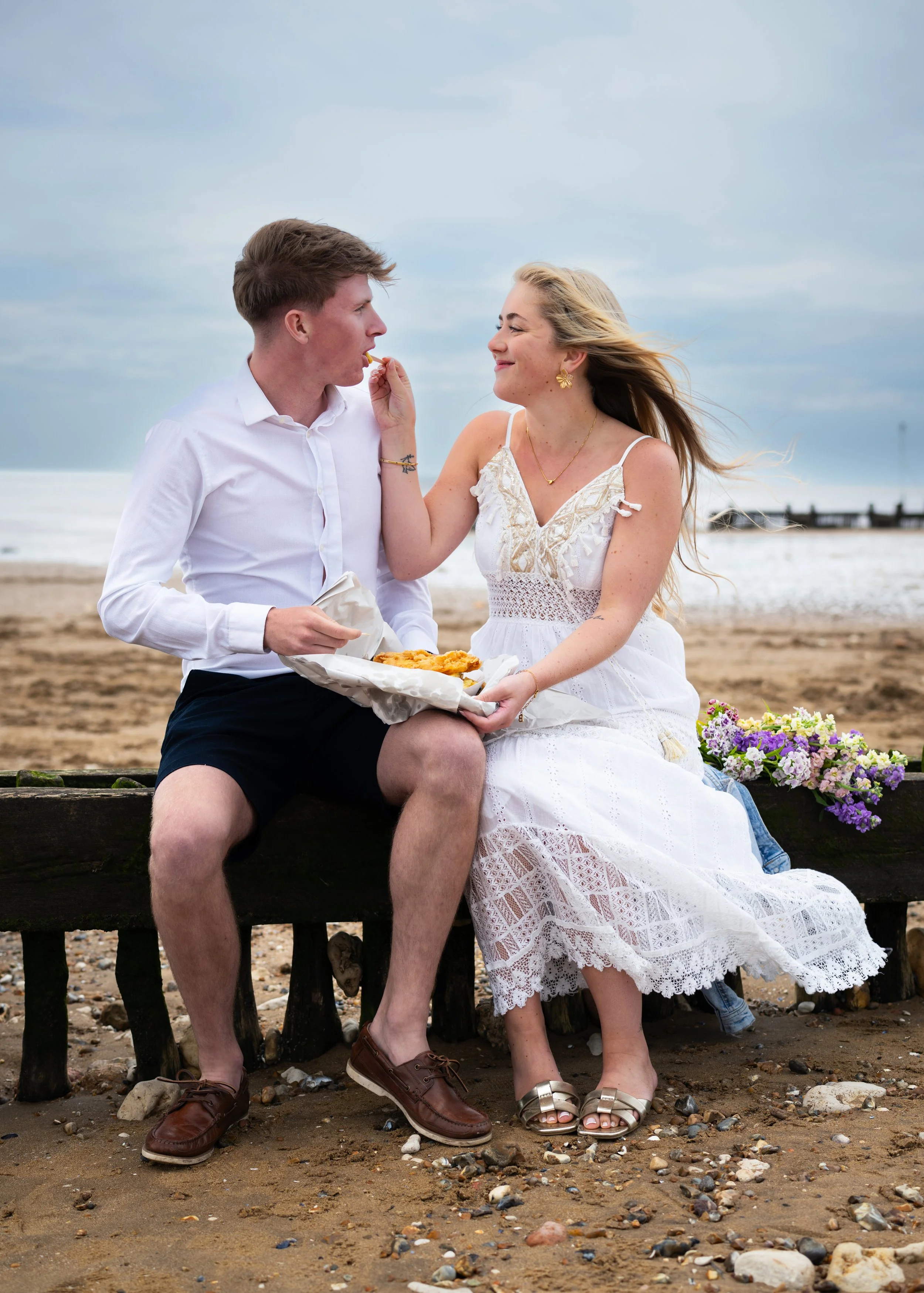 A young couple sits on a wooden bench at the beach. The man, wearing a white shirt and black shorts, is feeding the woman a piece of food. The woman, dressed in a white dress with lace, smiles at him. The background shows the ocean, a cloudy sky, and