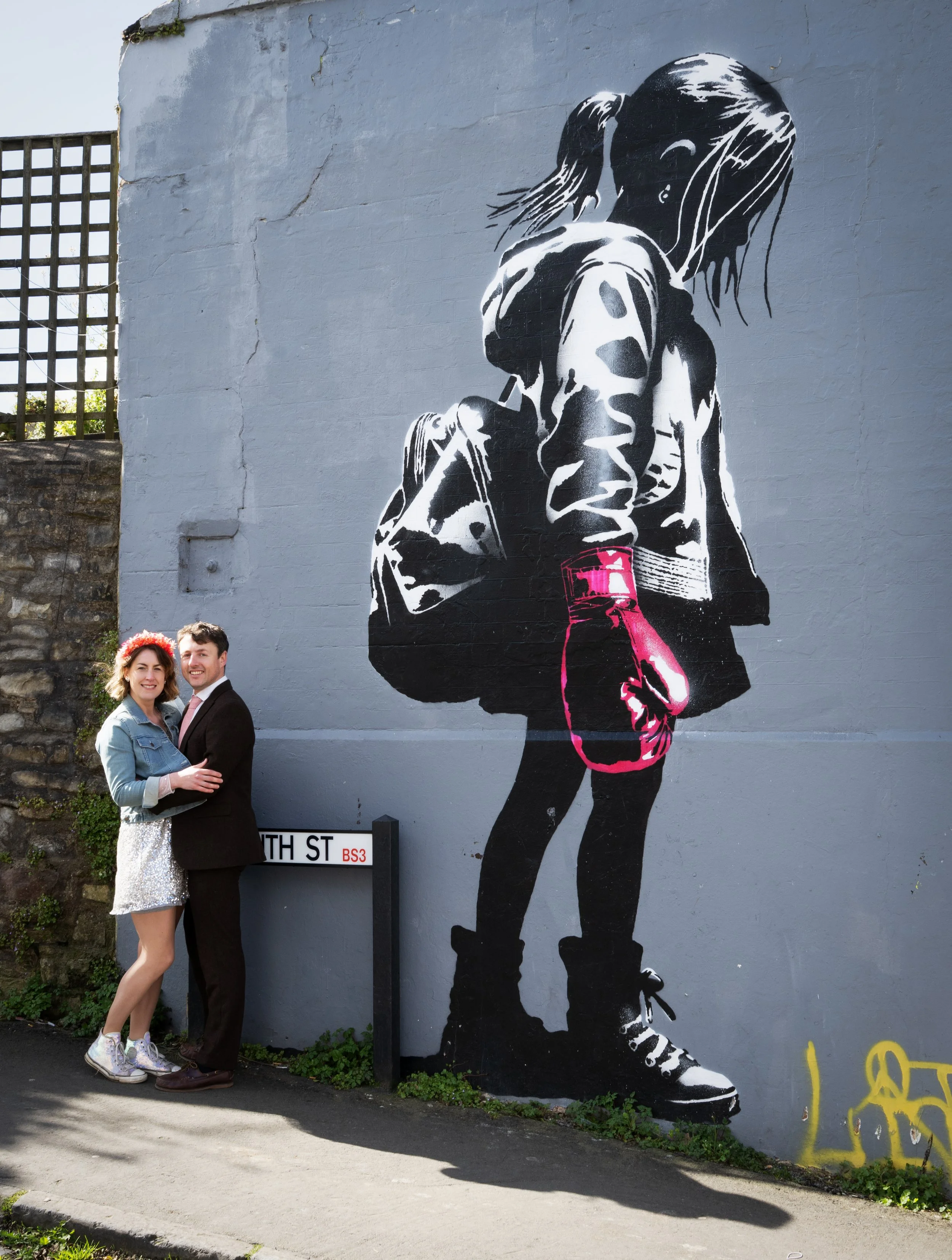 A couple standing close together, smiling, in front of a large street art mural of a girl with a backpack. The mural depicts the girl wearing a jacket, shorts, and sneakers, with a pink boxing glove on her hand, painted on a gray brick wall. There is