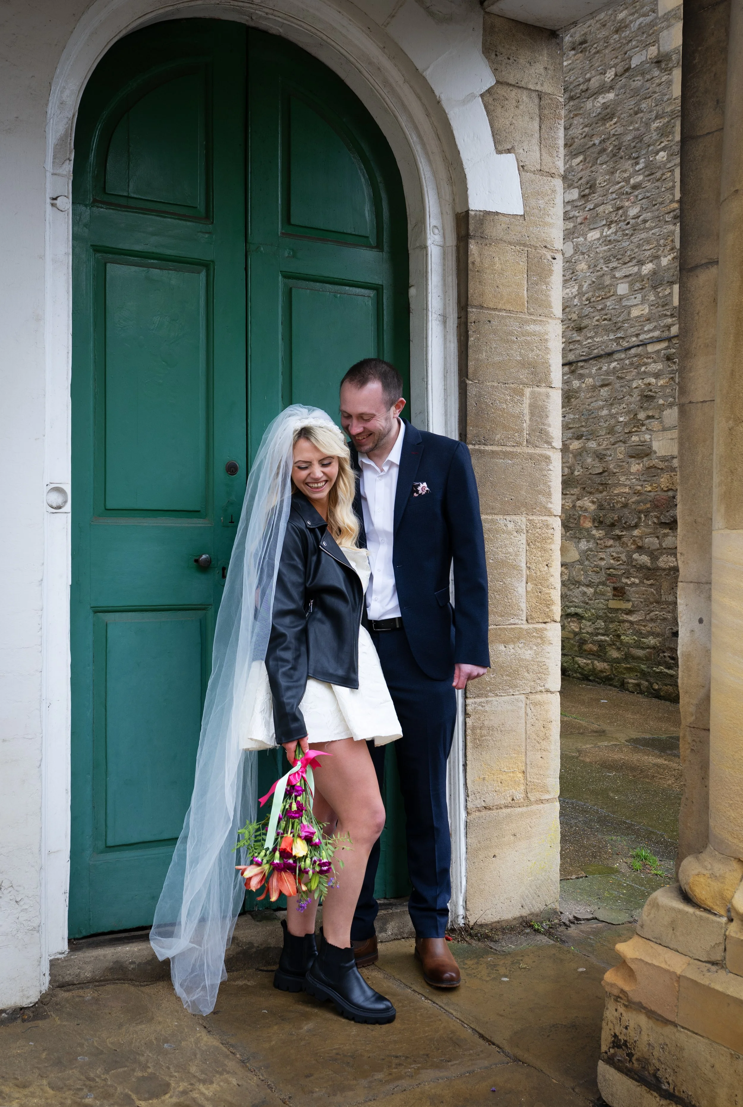 A smiling woman in a white dress and black boots holding a colorful bouquet, standing next to a smiling man in a suit, both leaning on a large green door at an old stone building. Wedding couple. Bride and Groom