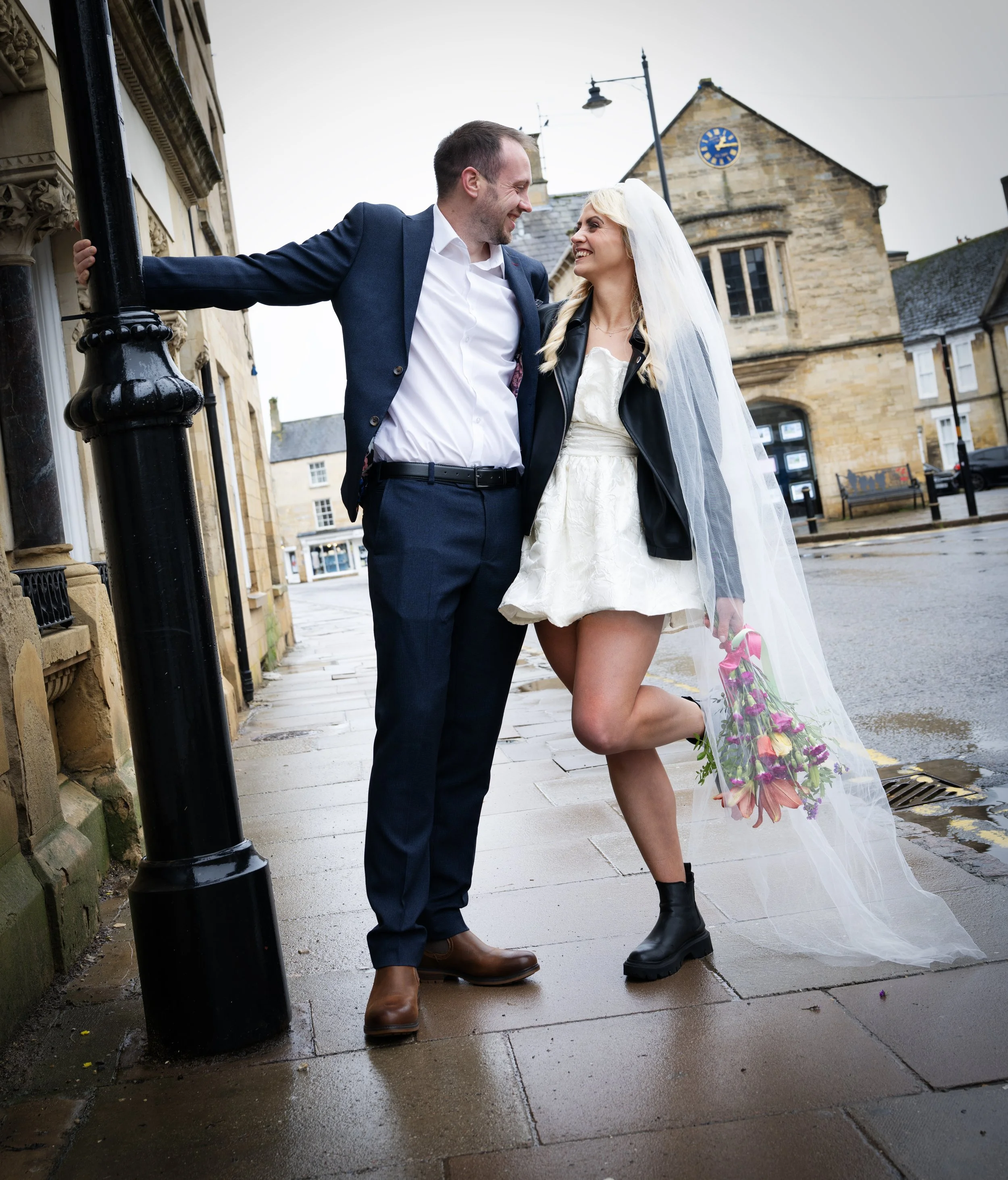 A newly married couple standing on a wet city sidewalk. The man is in a navy suit, leaning against a black lamppost, smiling at the woman. The woman wears a white dress, combat boots, holds a bouquet, and has a long veil. They are looking at each oth