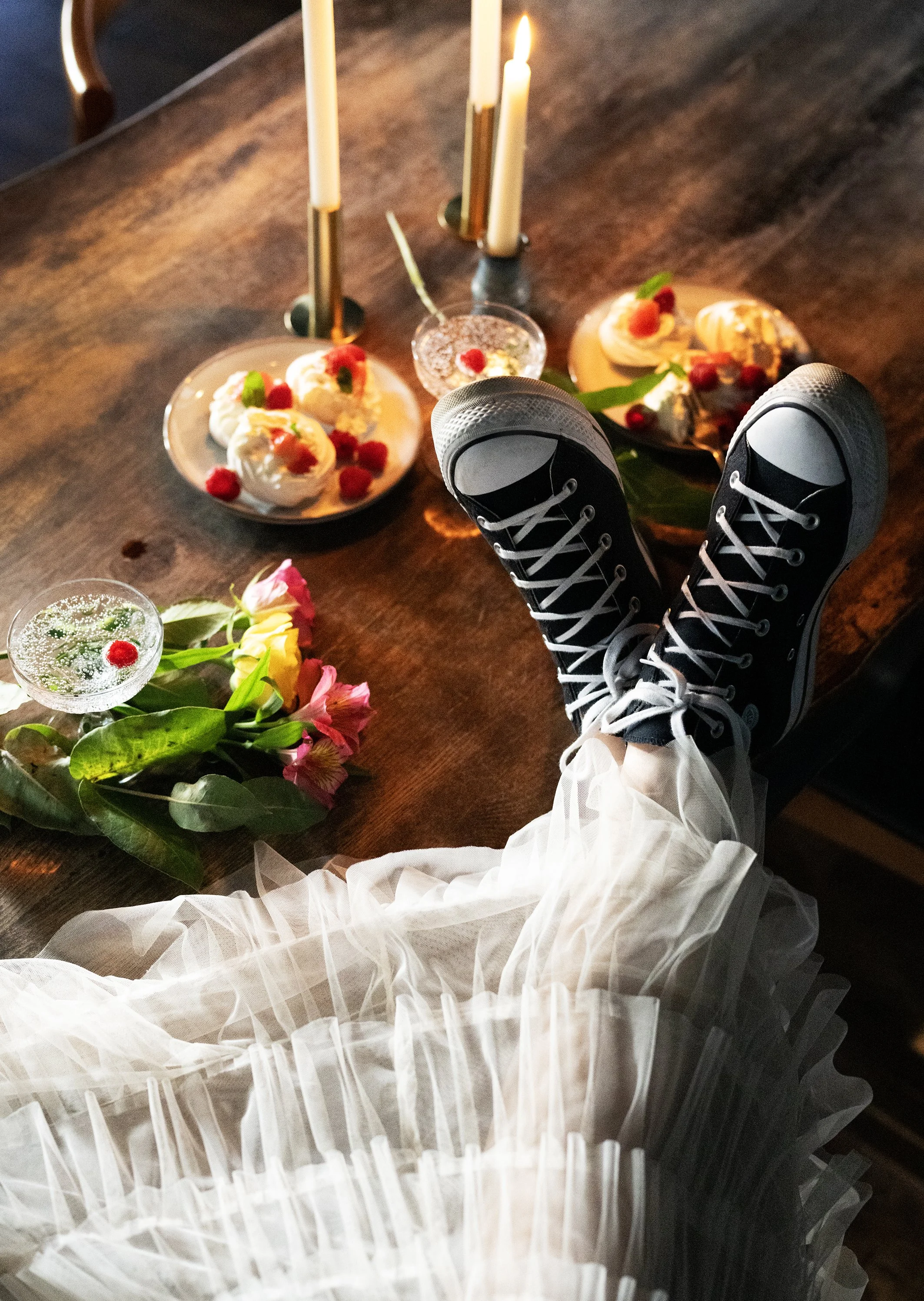 Bride at a table with converse on and a white tulle skirt. On the table, there are two plates with three desserts topped with red berries and garnishes, lit candles, a cocktail glass, and a bouquet of pink and white flowers.