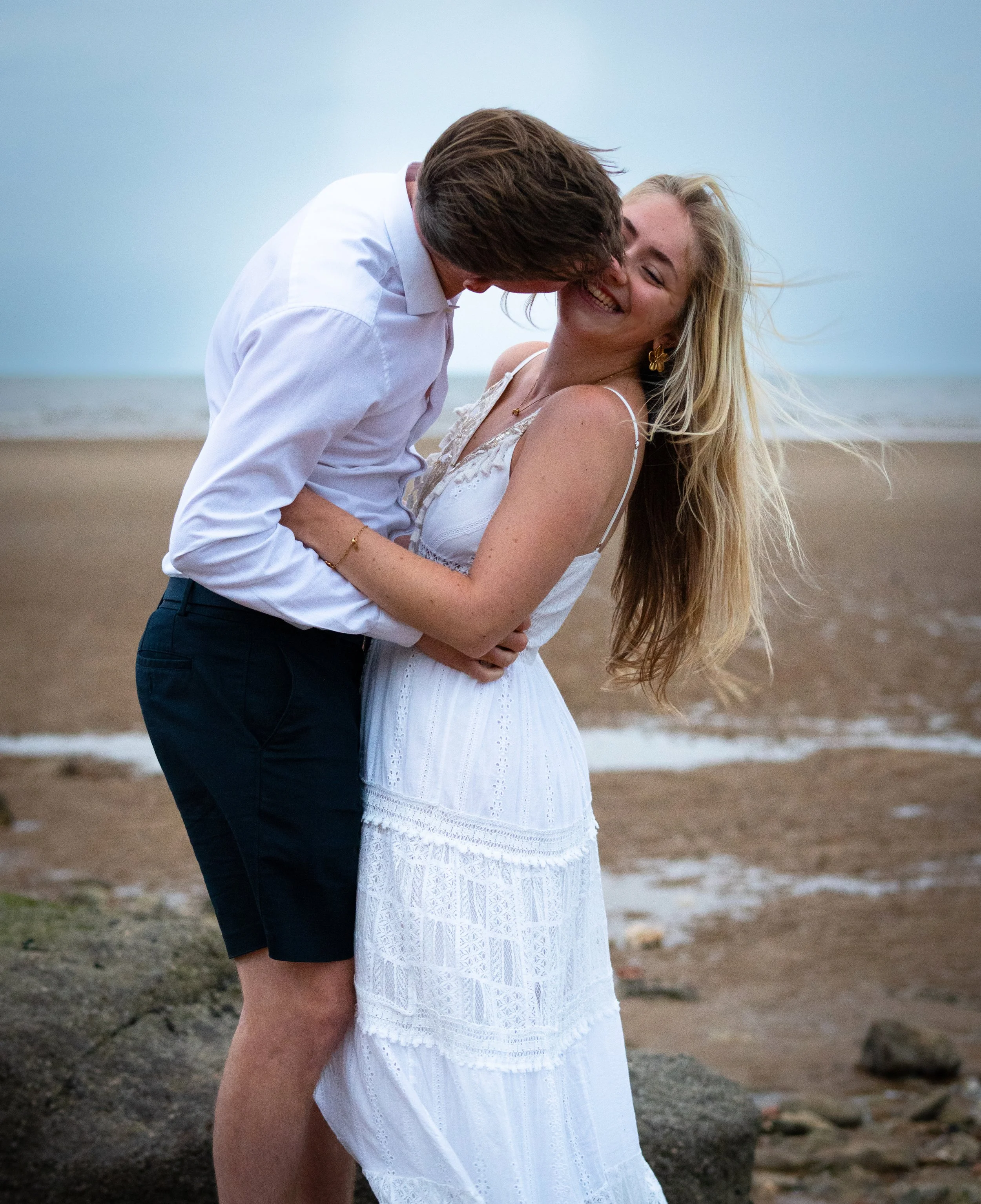 A young man and woman embrace and kiss on a beach, with the ocean and sandy shoreline in the background.