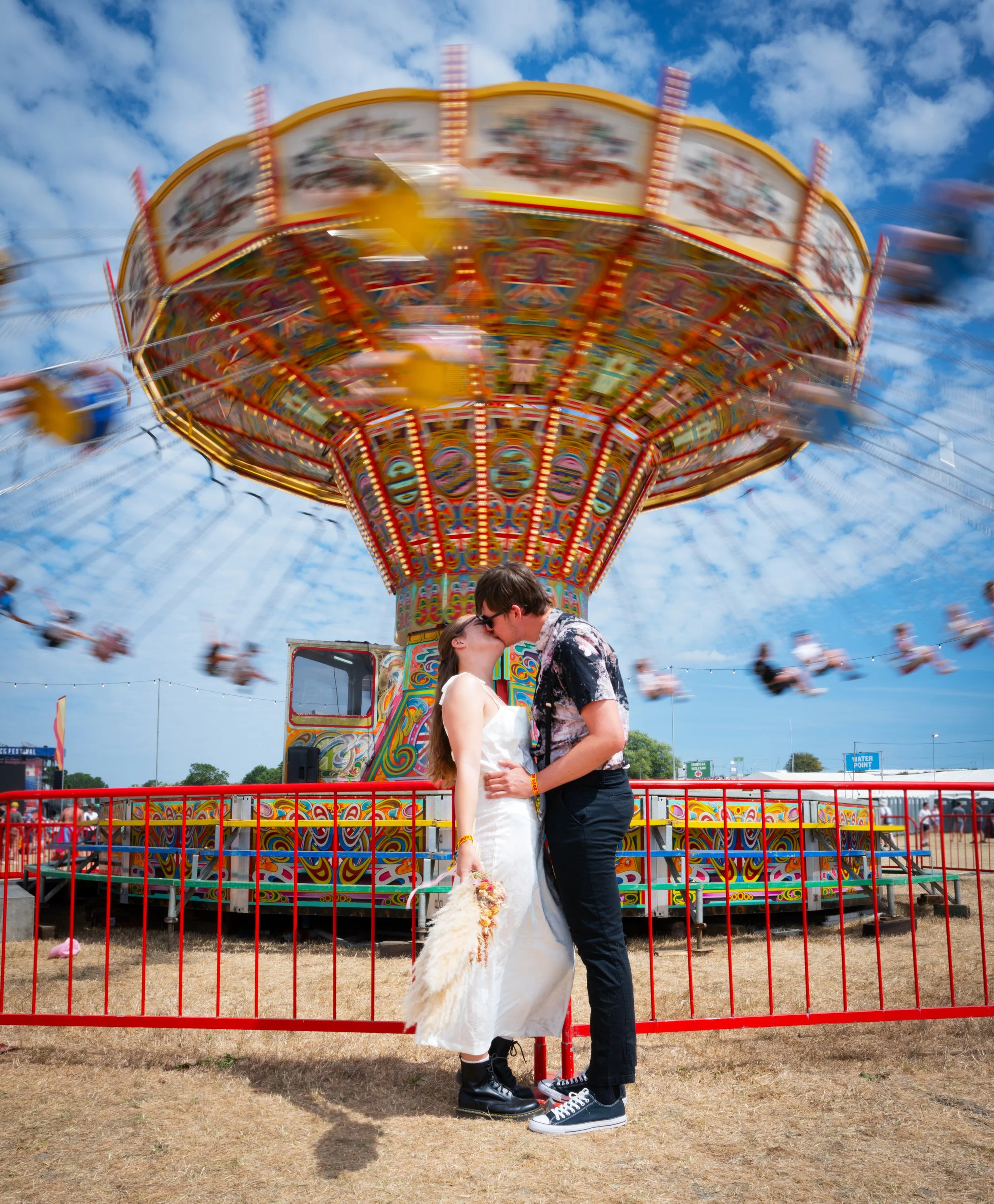 Bride and groom kissing in front of a colourful spinning swing ride at a festival-style wedding.