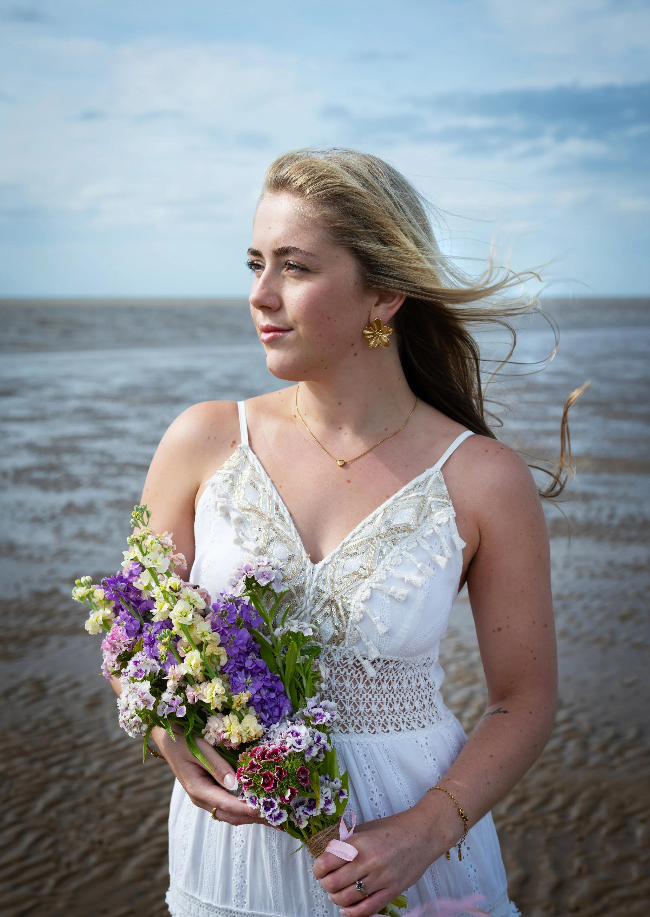 A woman with blond hair and a white dress holding a bouquet of purple and white flowers on a beach with a cloudy sky.