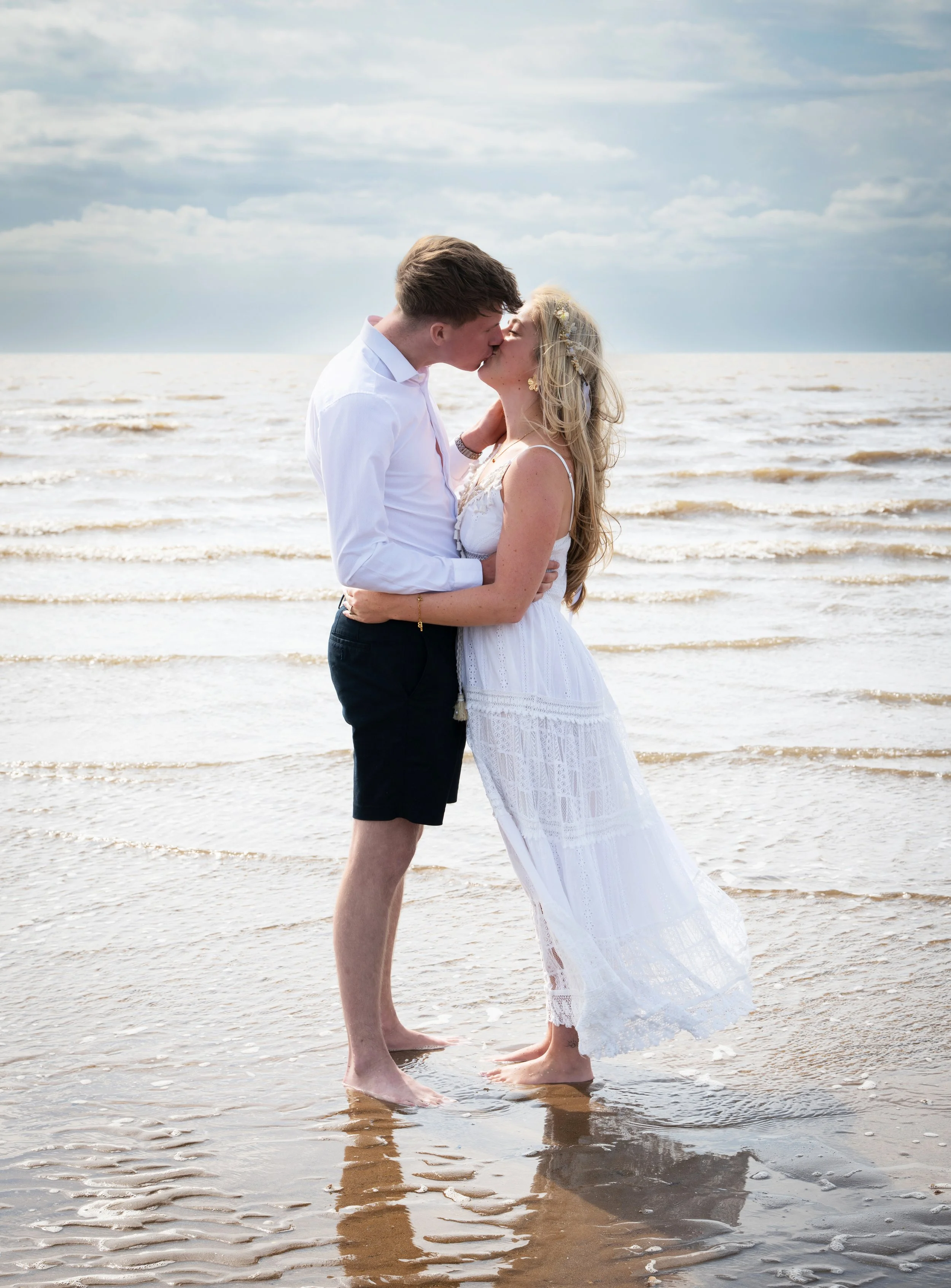 A couple kissing on the beach with ocean waves, cloudy sky, and sandy shoreline.