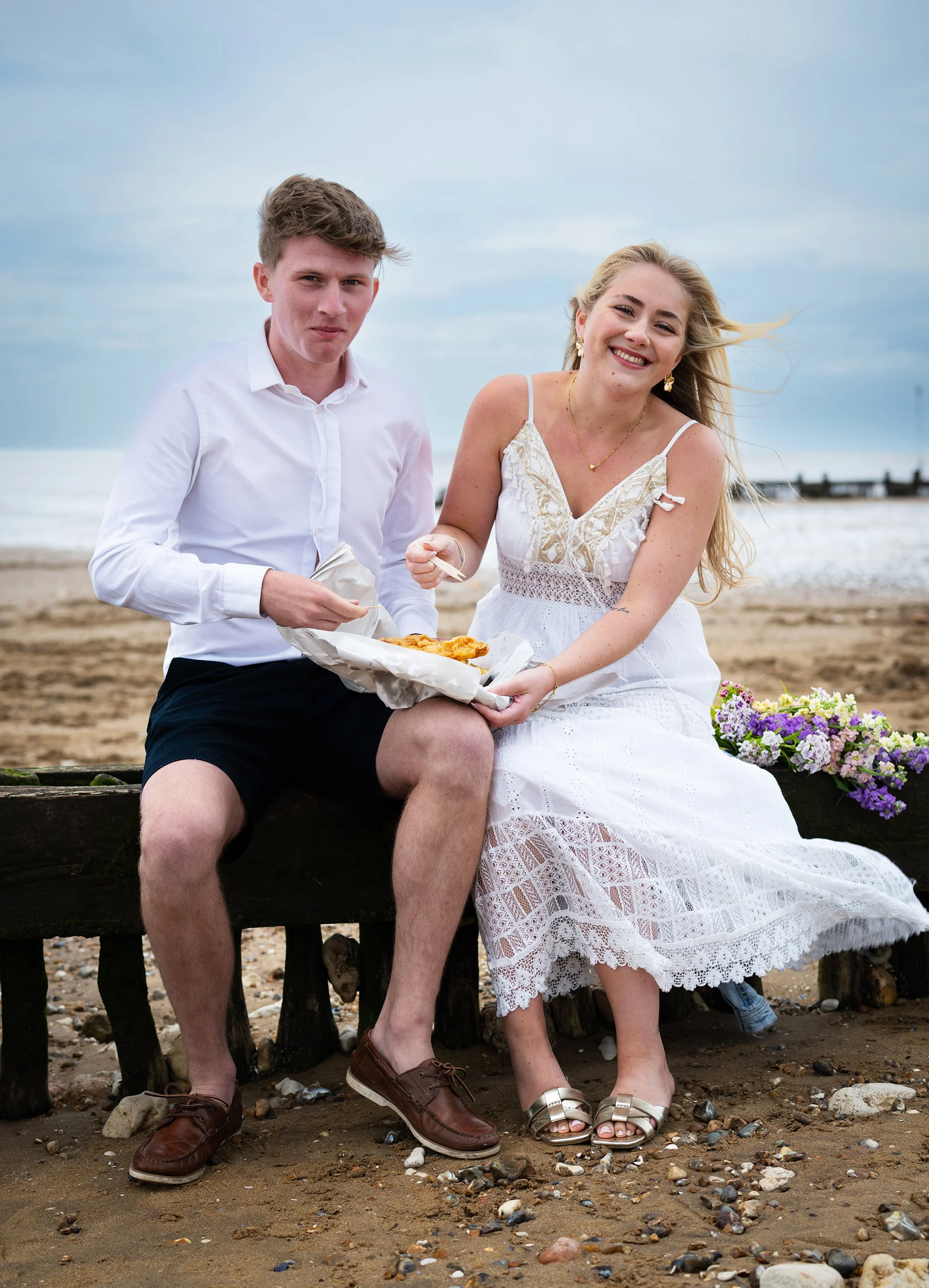 Newly married couple eating fish and chips together on the beach.