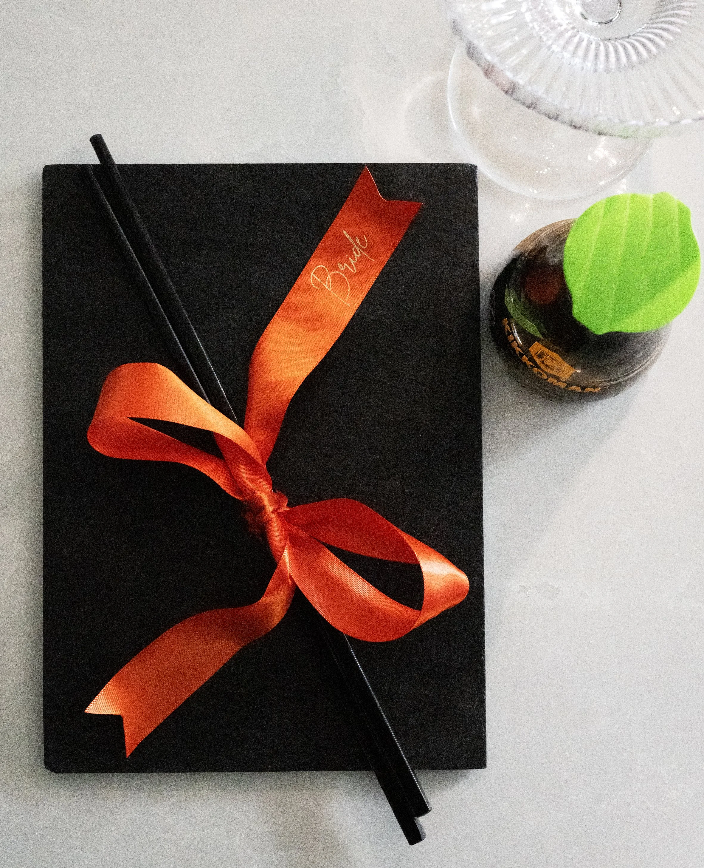 A black gift box decorated with an orange ribbon tied in a bow and a small orange tag that says 'Bride'. The box is set on a white surface next to black chopsticks, a clear glass vase, and a dark bottle with a green lid. Wedding breakfast sushi