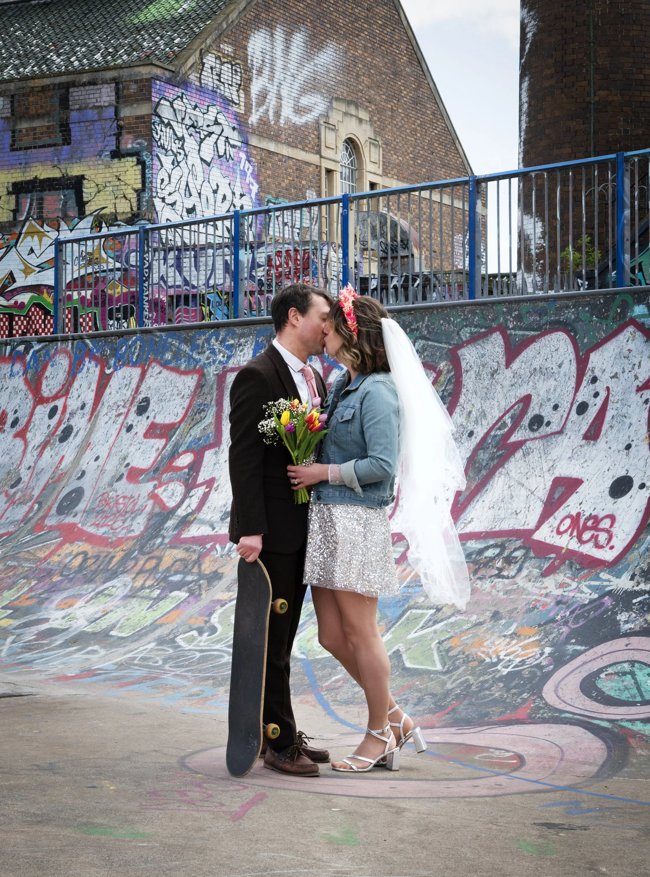 A wedding couple in wedding attire sharing a kiss in a graffiti-covered skate park, with the groom holding a skateboard and the bride holding a bouquet of flowers. Bristol Dean lane Skatepark