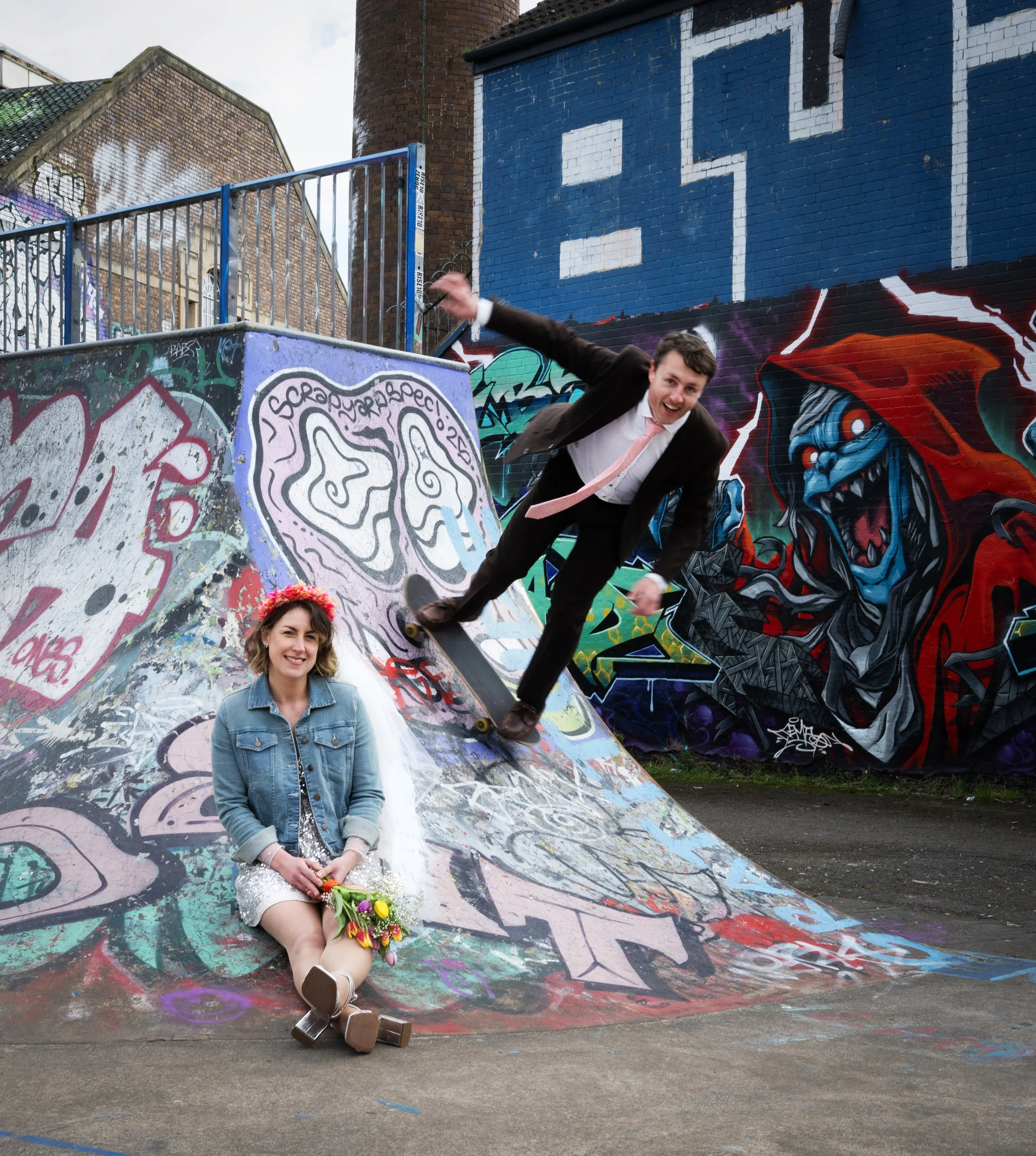 A man in a suit skateboarding on graffiti-covered ramp with a woman dressed as a bride sitting below holding flowers, in front of a colorful graffiti mural featuring a blue-faced monstrous figure with red hood.