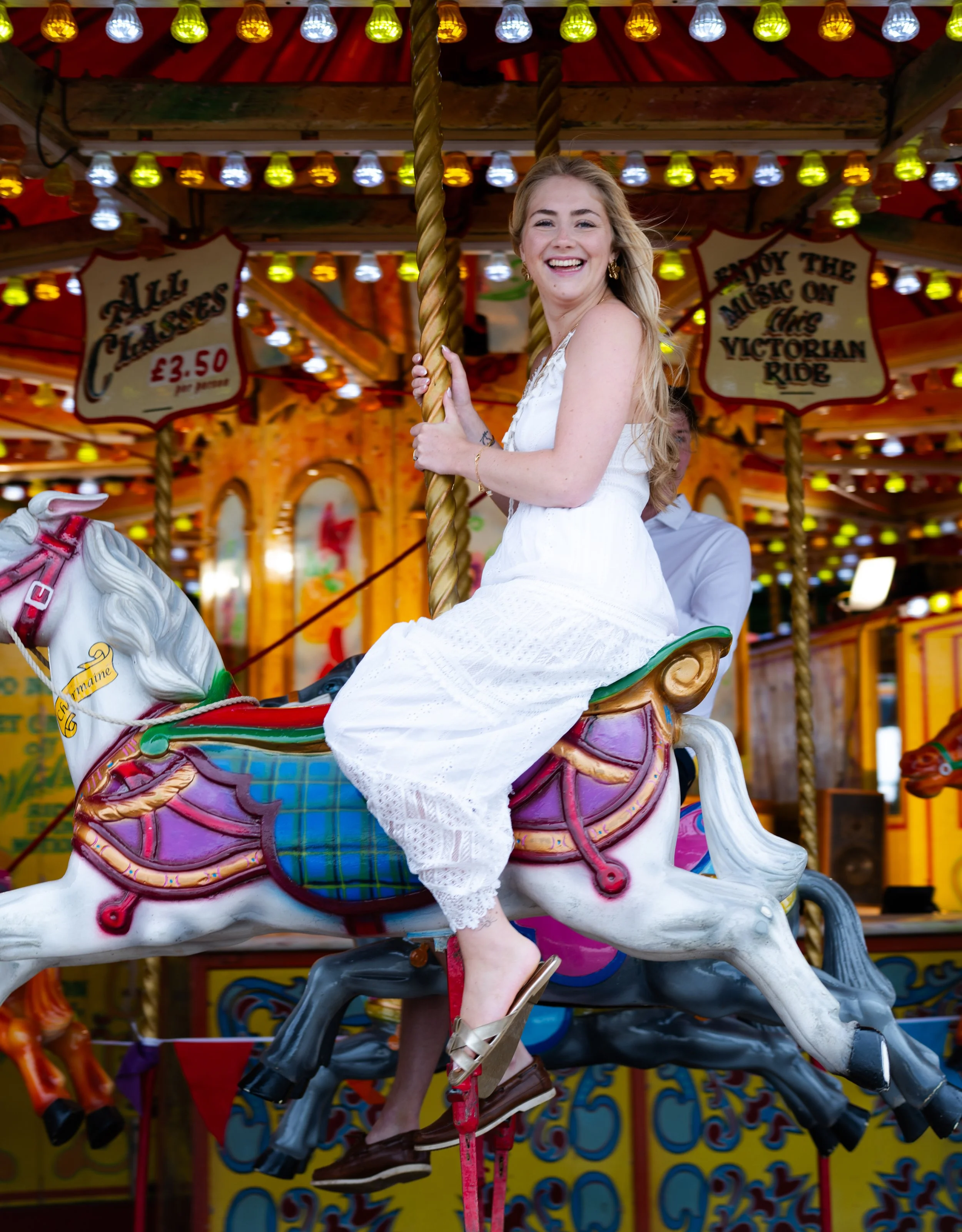 A woman riding a carousel horse at a fair, smiling and holding onto the pole. The carousel is decorated with colorful lights and signs, including one that says 'Enjoy the music on this Victorian ride' and another that states the cost as £3.50 per per