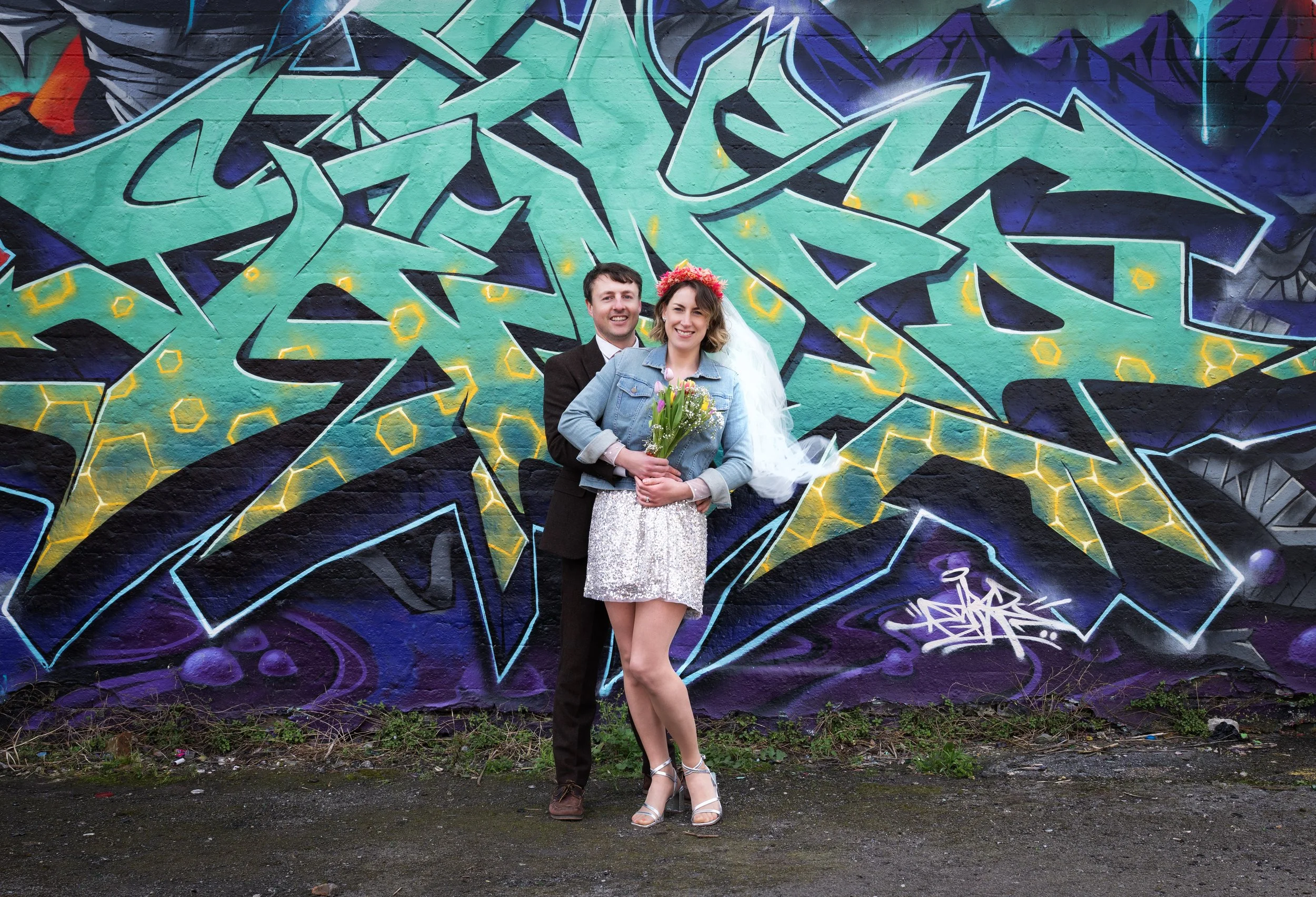 A smiling couple, a woman holding a bouquet of flowers, stand in front of colorful graffiti art on a brick wall. The woman wears a denim jacket, a sparkly skirt, and heels, with a flower crown, while the man wears a dark jacket. Dean lane Skatepark