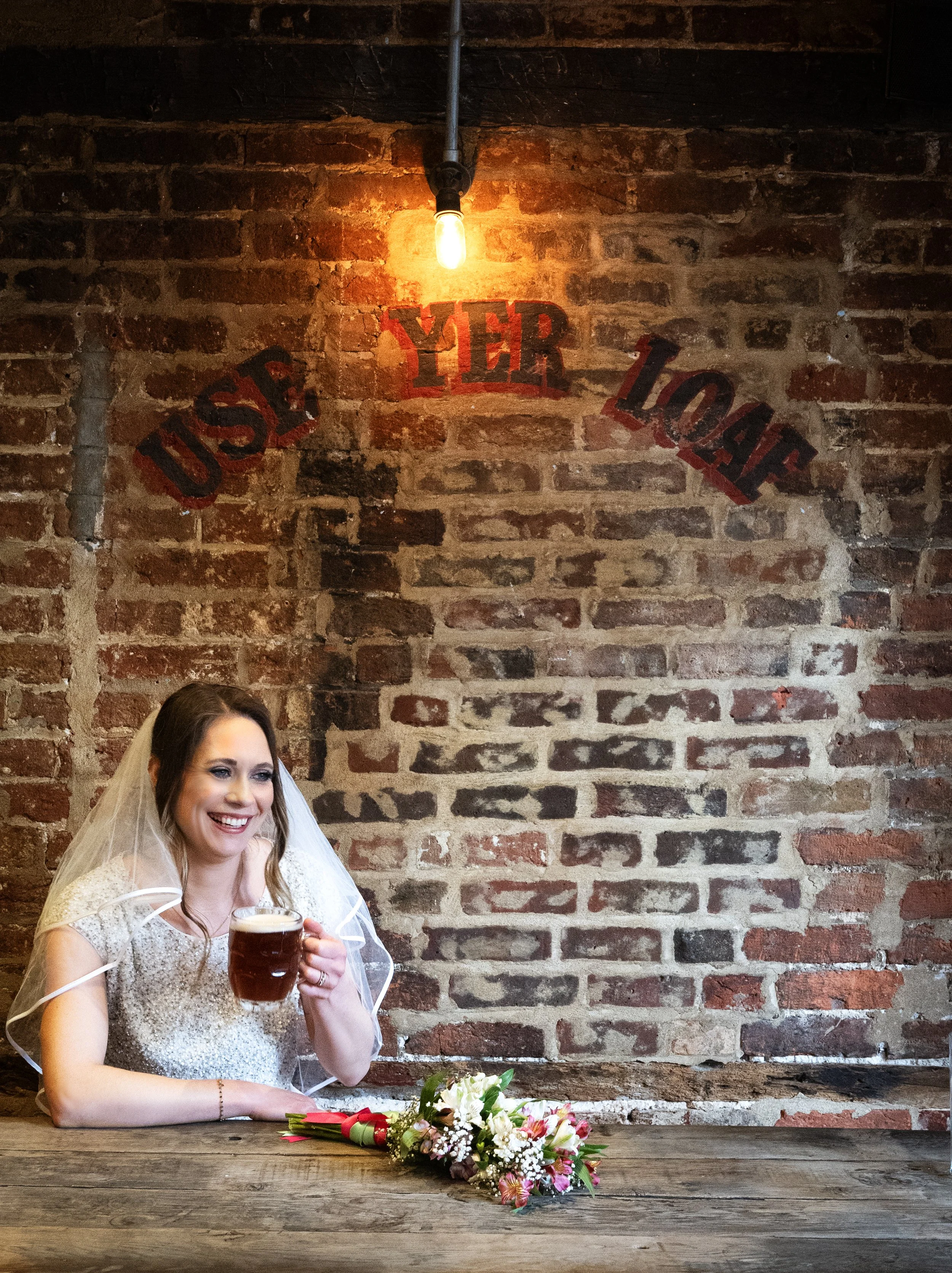 A smiling bride sitting at a rustic wooden table with a flower bouquet, holding a mug of beer, against a brick wall background with the words 'Use your loaf' painted on it and a hanging light bulb above. Bristol Christmas Steps