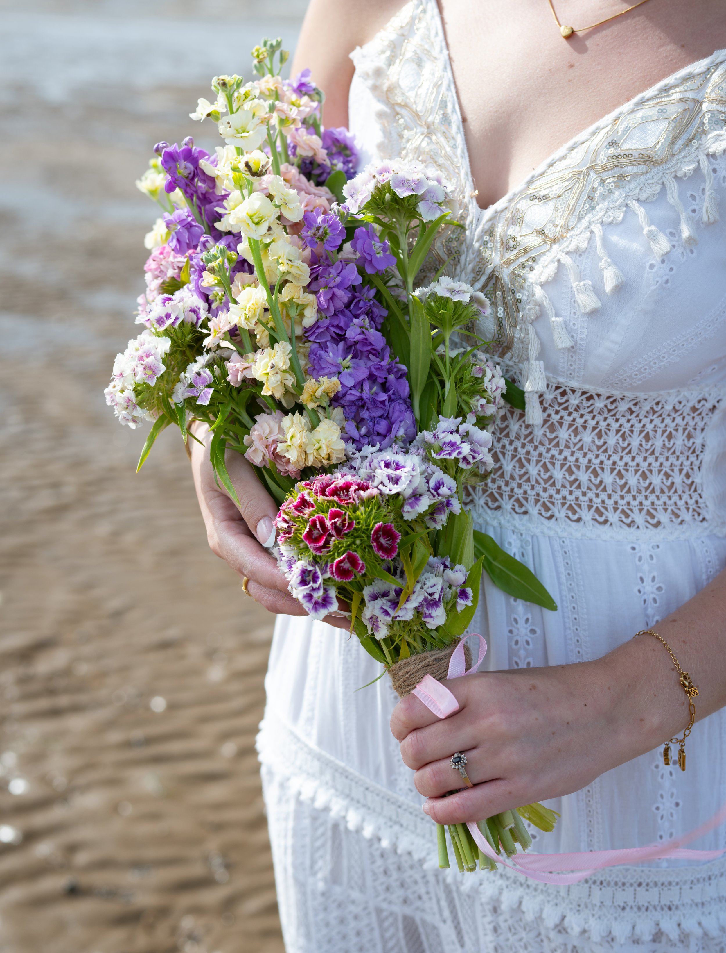A woman in a white dress holding a bouquet of purple, white, and pink flowers at the beach.
